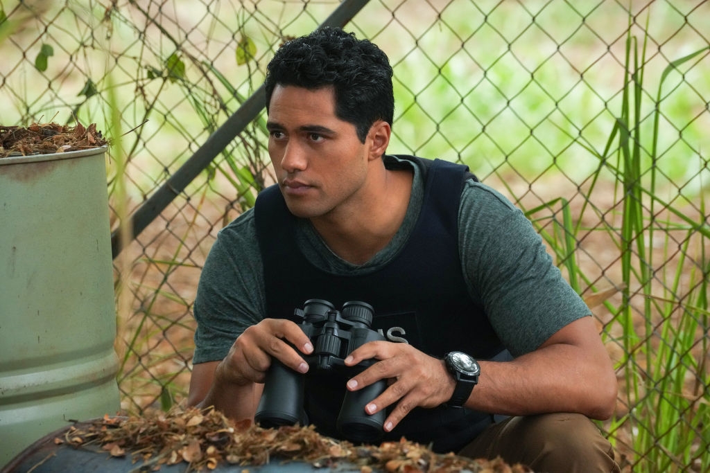 Person kneeling outdoors, wearing a tactical vest and holding binoculars, appears focused. Chain-link fence and foliage in the background