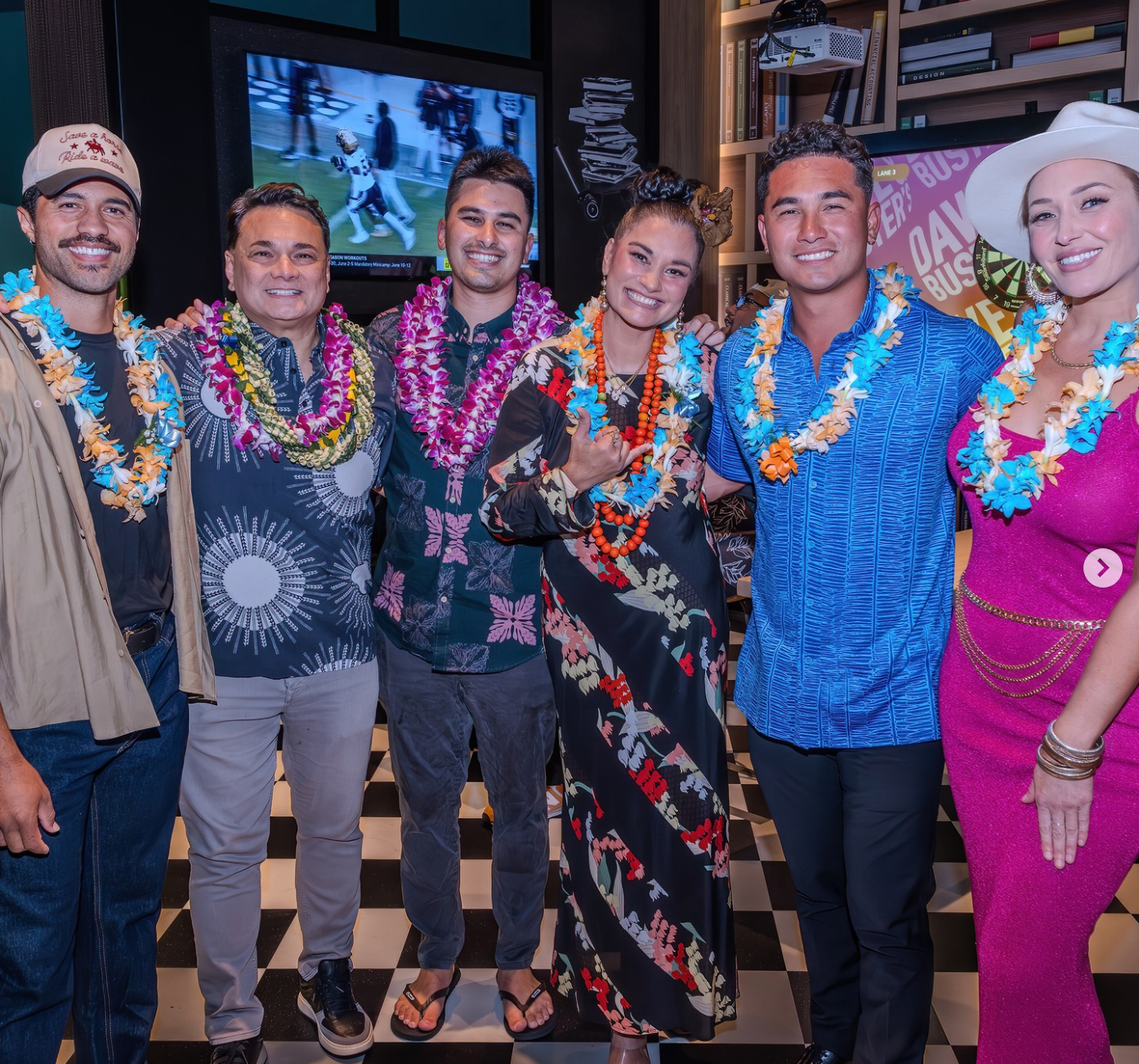 Six people wearing leis pose together, some in floral or patterned outfits, looking joyful at an event indoors