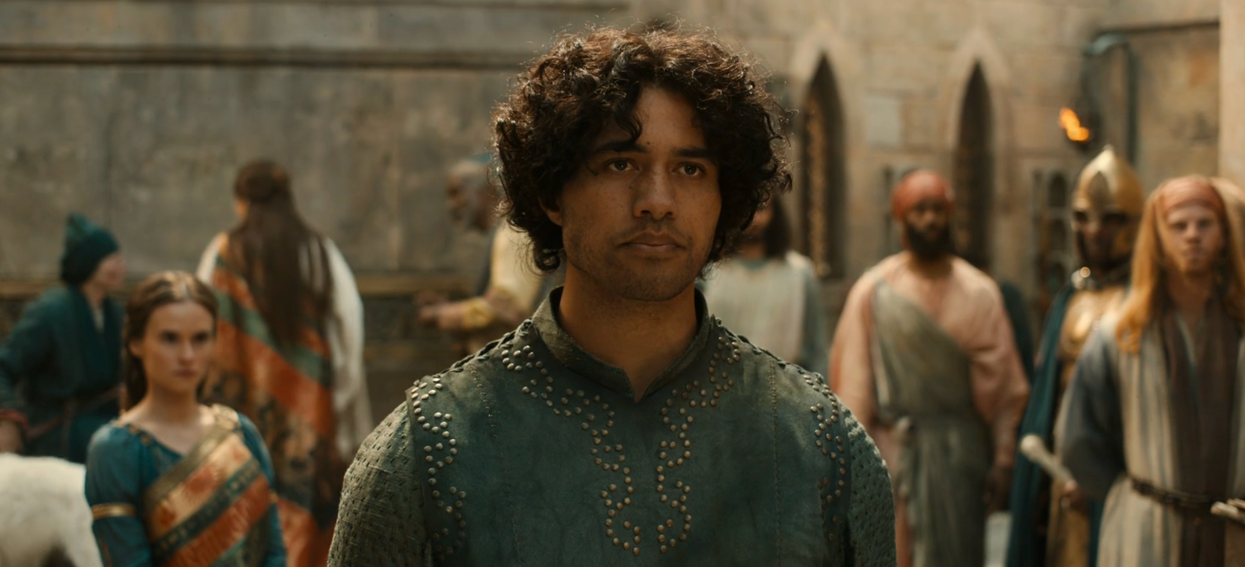 A person with curly hair in patterned attire stands in a medieval-style setting, surrounded by people in historical costumes