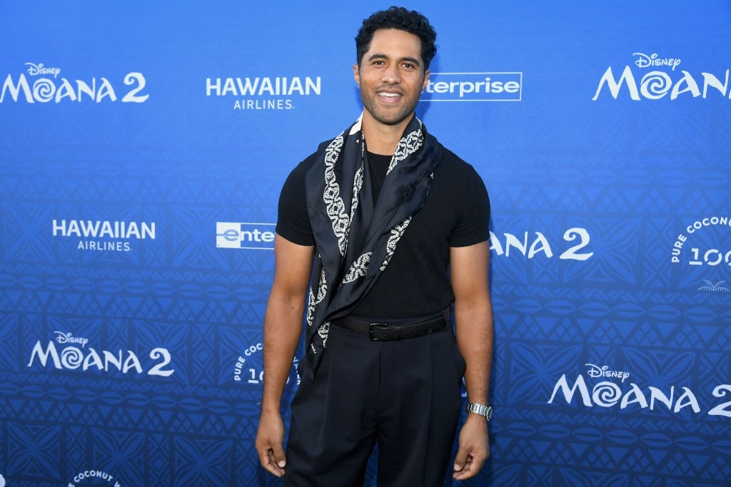 Man in a black shirt and slacks with a patterned scarf poses on the red carpet for a "Moana 2" event backdrop