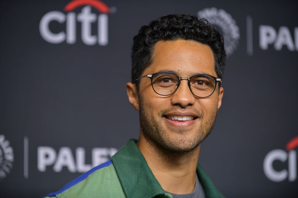 A person with short hair and glasses smiles at an event, wearing a casual jacket over a t-shirt