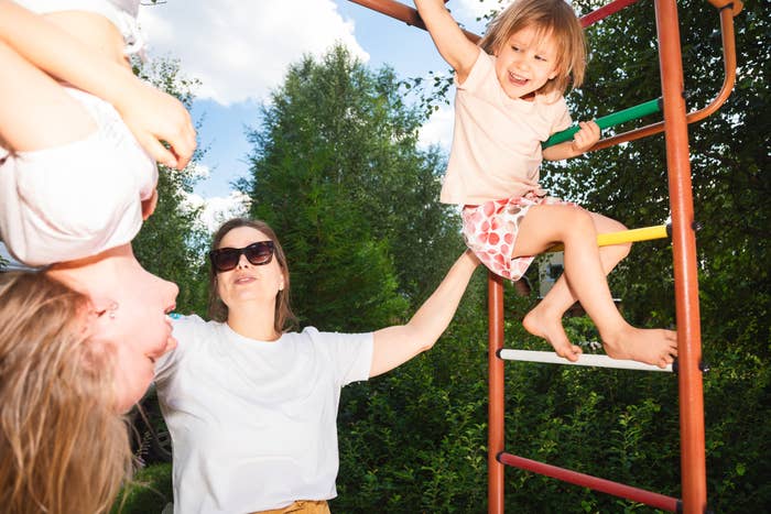A parent holds a child hanging upside down and smiles at another child sitting on a jungle gym, outdoors