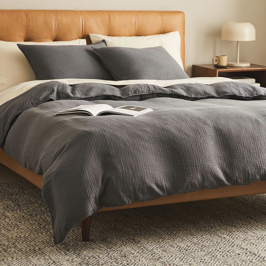 Elegant bedroom with a wooden bed frame, gray bedding, and an open book. A nightstand holds a lamp and a mug. Cozy and stylish for shopping inspiration