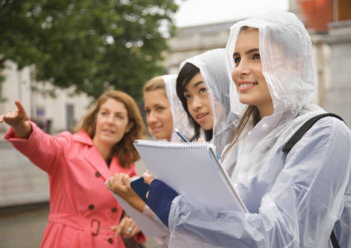 Four women outdoors wearing rain ponchos, one pointing, others holding notebooks, appearing to observe or study something off-camera