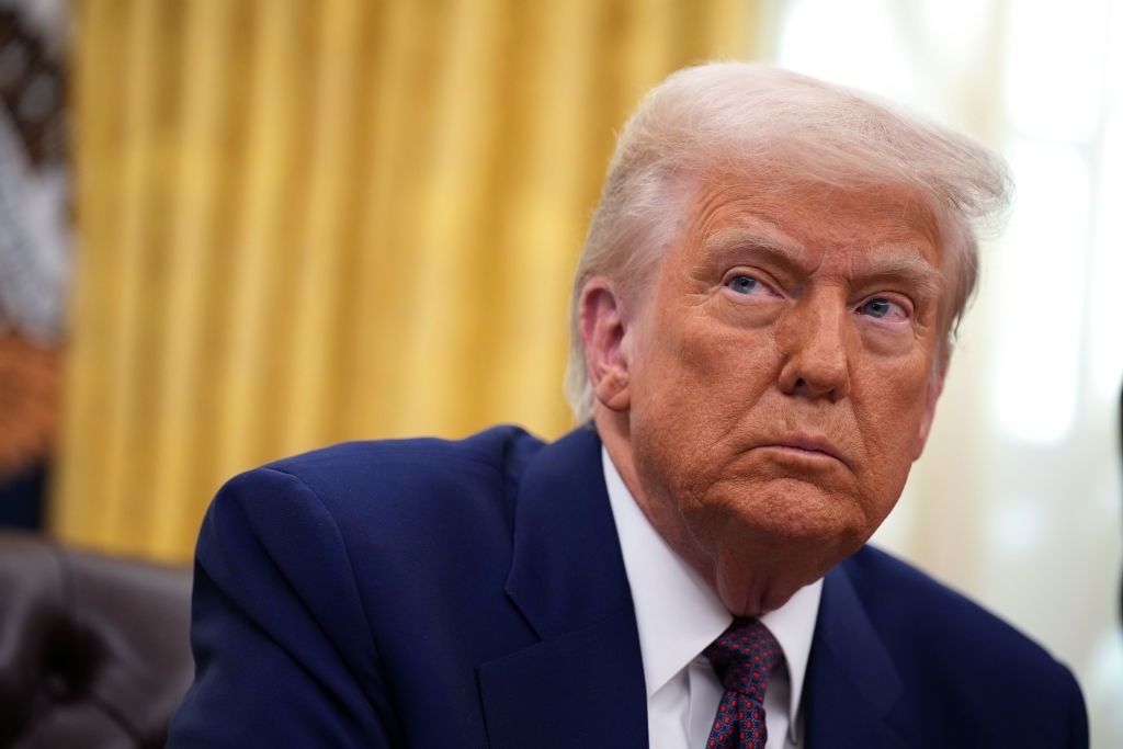 A person in a suit sits in an office, looking off-camera with a serious expression