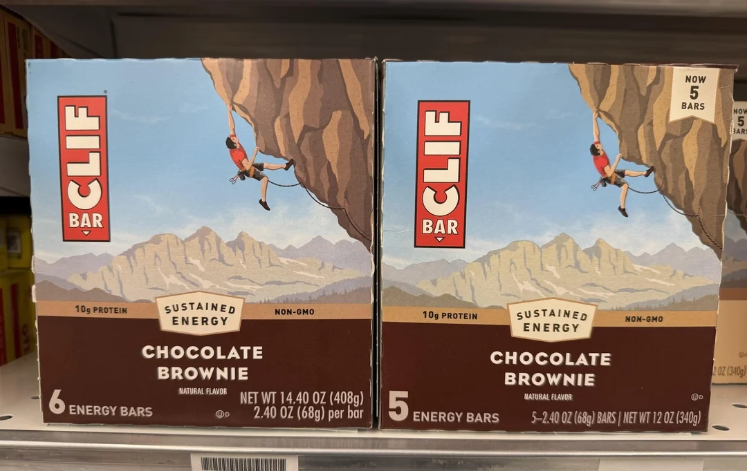 Two boxes of Clif Bar chocolate brownie energy bars on a store shelf, one containing 6 bars and the other containing 5 with a call out on the box that says "now 5 bars"