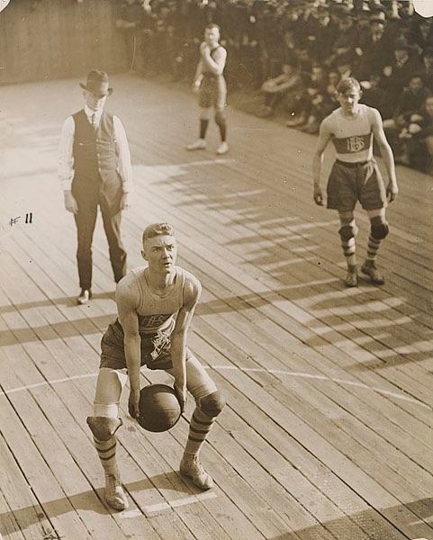 Vintage photo of a basketball player preparing to shoot a free throw, with other players and a referee in early 20th-century attire