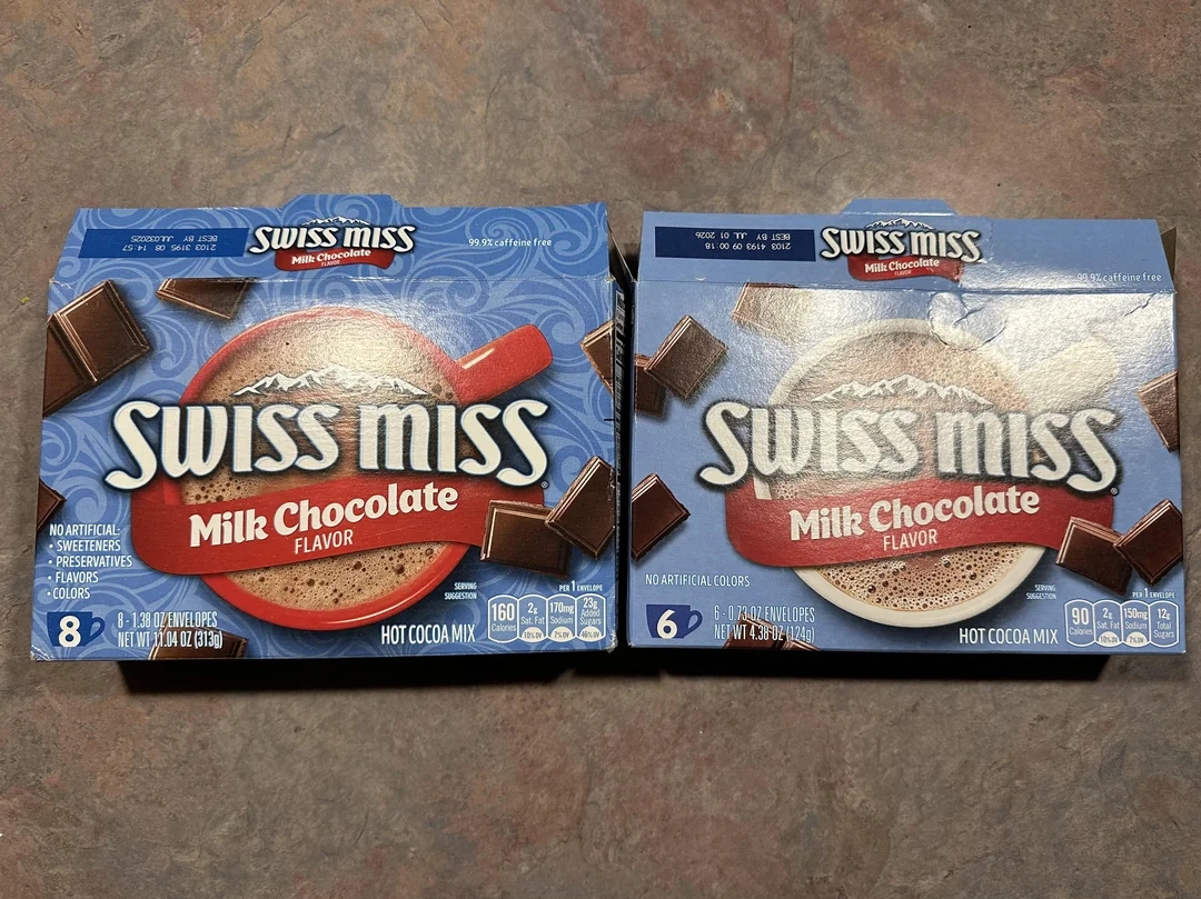 Two Swiss Miss Milk Chocolate hot cocoa mix boxes, one containing 8 packets and the other 6 packets, on a textured surface