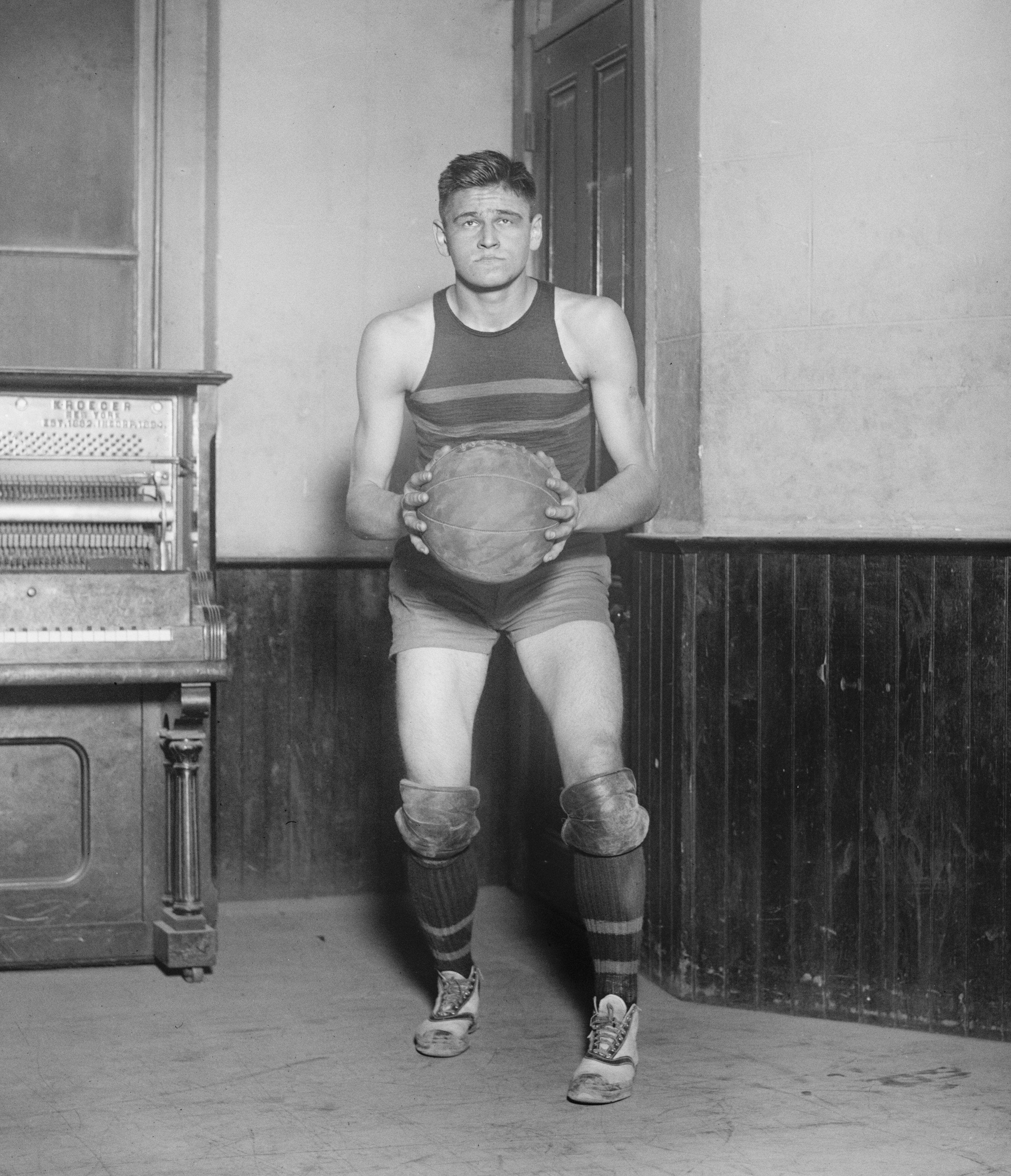 A vintage basketball player in a 1920s-style athletic outfit holds a basketball indoors, standing near a piano