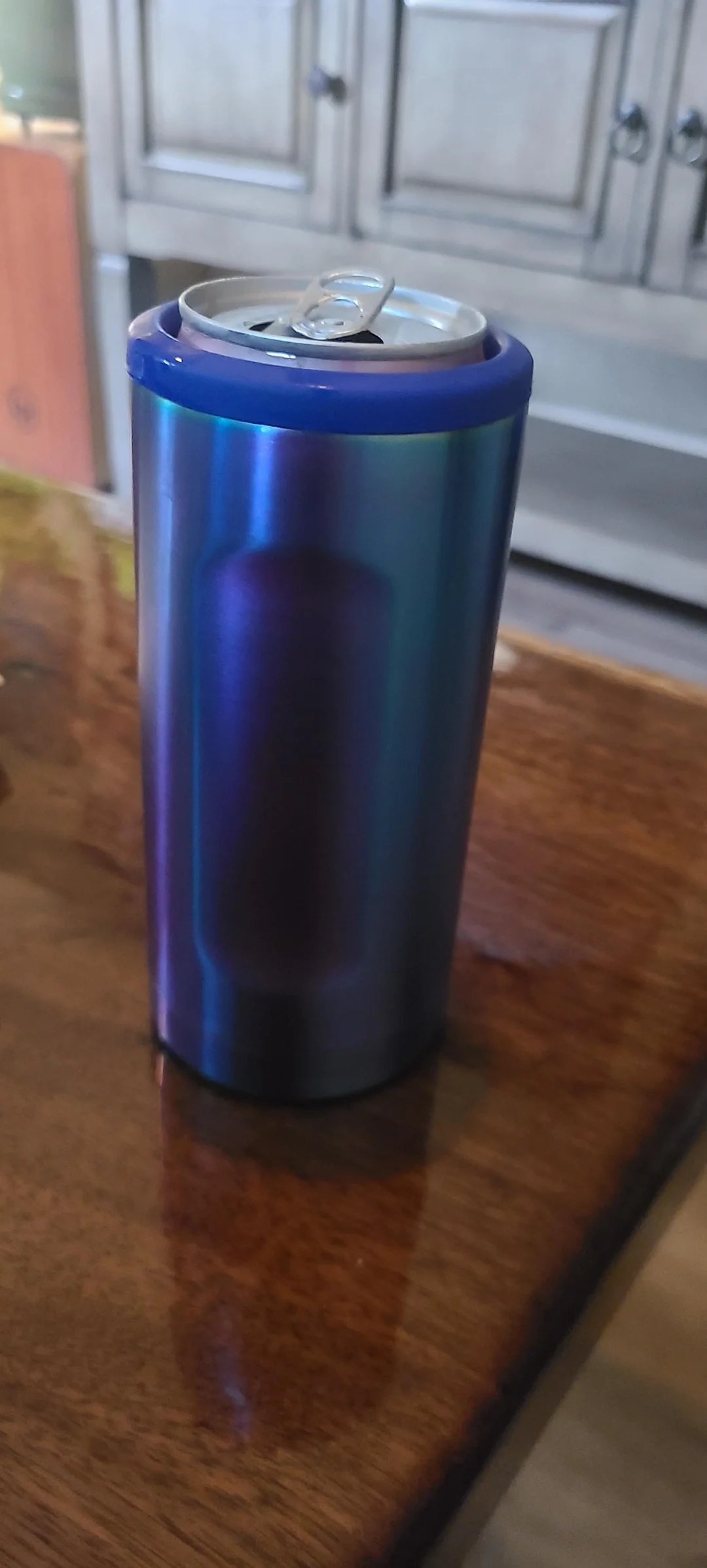 A drink can in a sleek holder sits on a wooden table. Background shows part of a cabinet