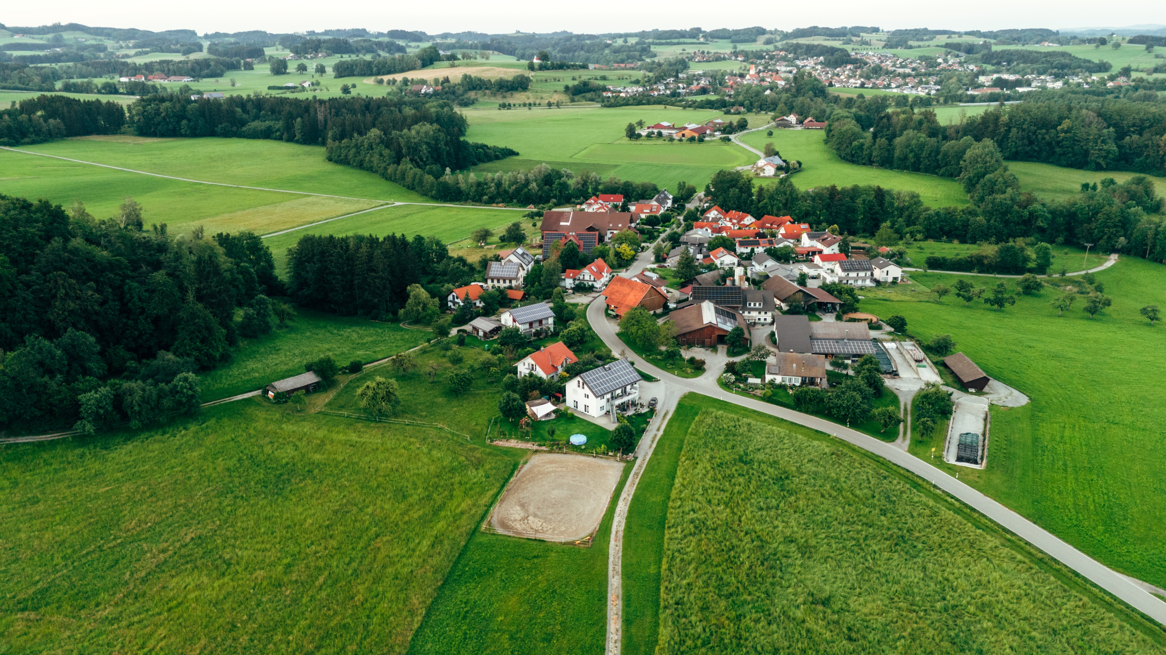 Aerial view of a small village surrounded by lush green fields and forests, with clusters of houses and a few roads intersecting the area