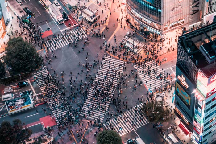 Aerial view of a bustling city intersection with numerous pedestrians crossing at various angles, surrounded by tall buildings and bright lights