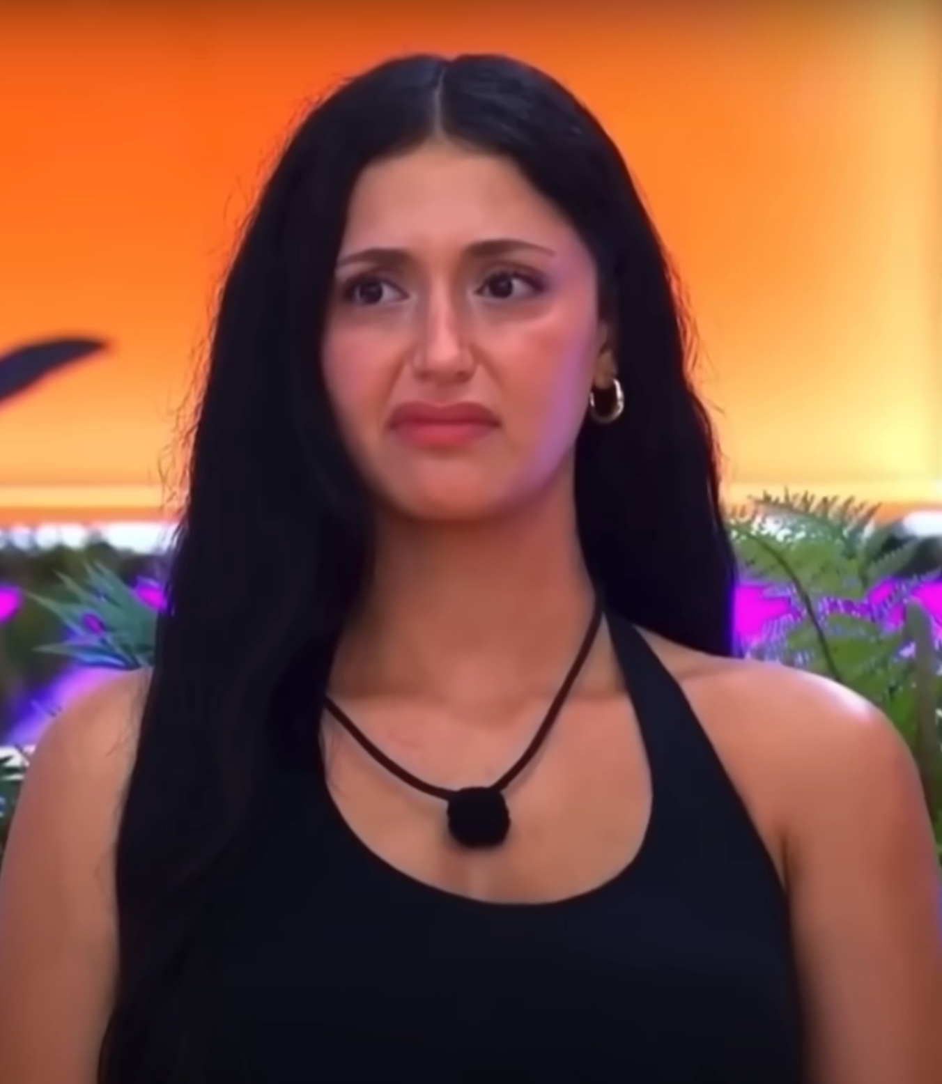 A person with long hair looks ahead with a skeptical expression, wearing a sleeveless top and hoop earrings