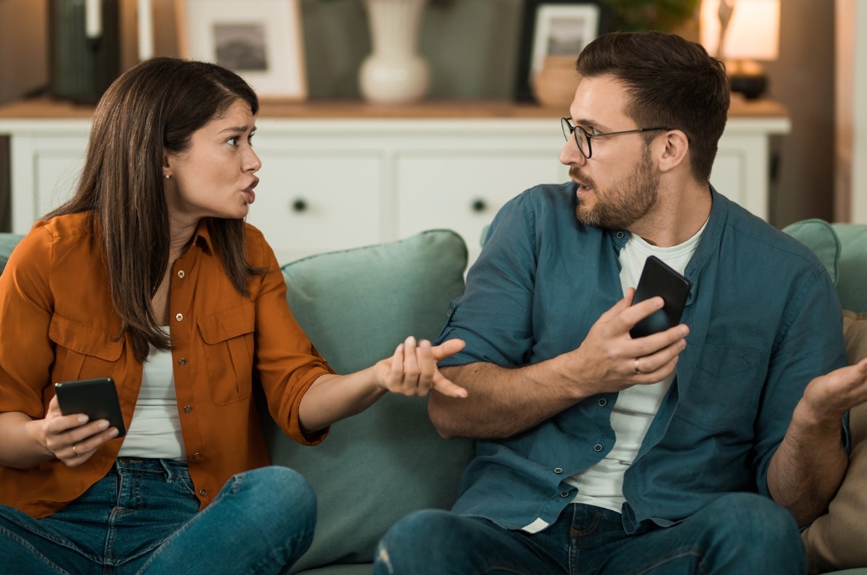 A couple sitting on a sofa in a heated discussion, holding smartphones, gesturing expressively at each other