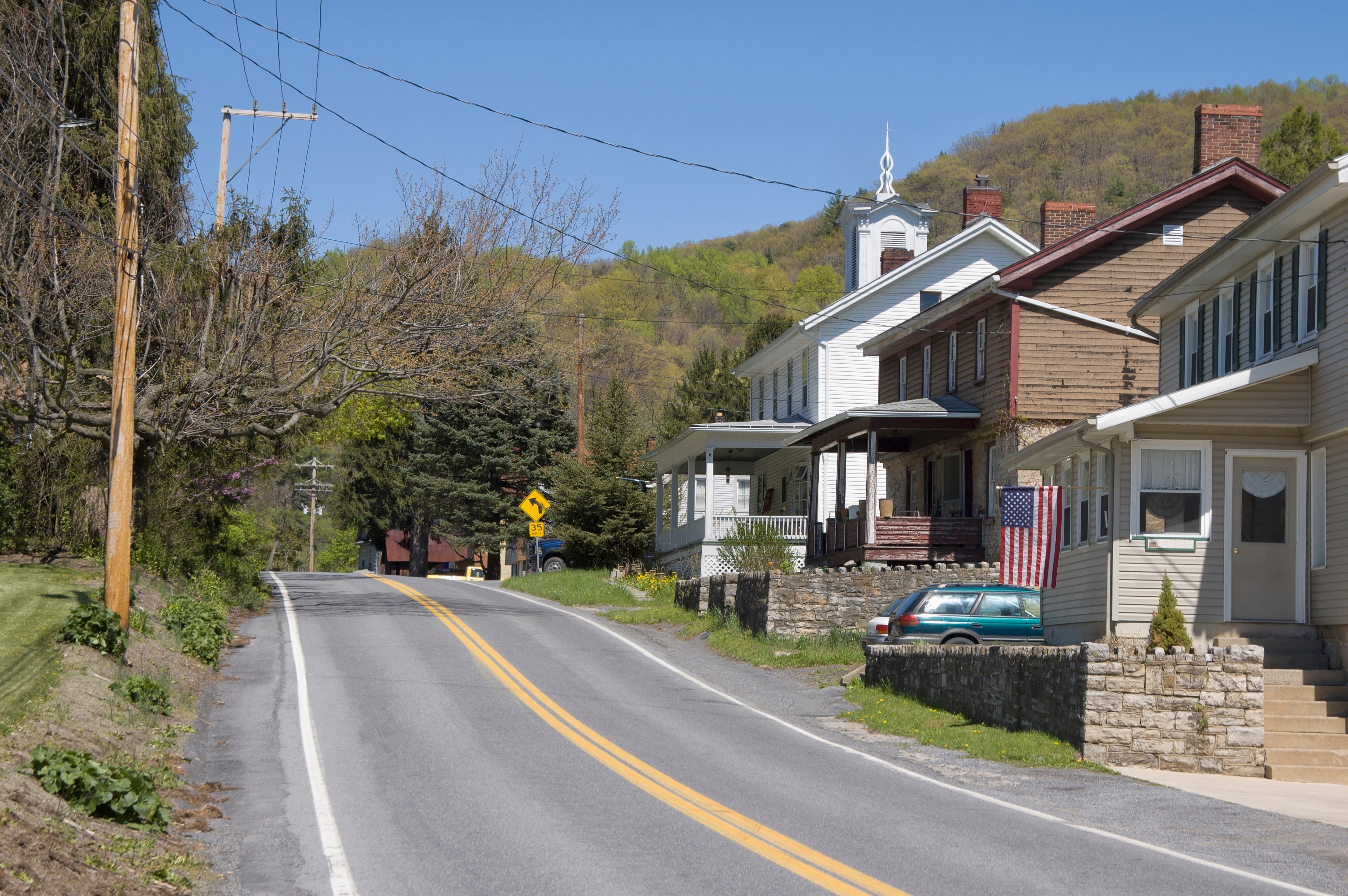 A quiet rural road lined with houses, a parked car, and distant hills under a clear sky. An American flag hangs from one house