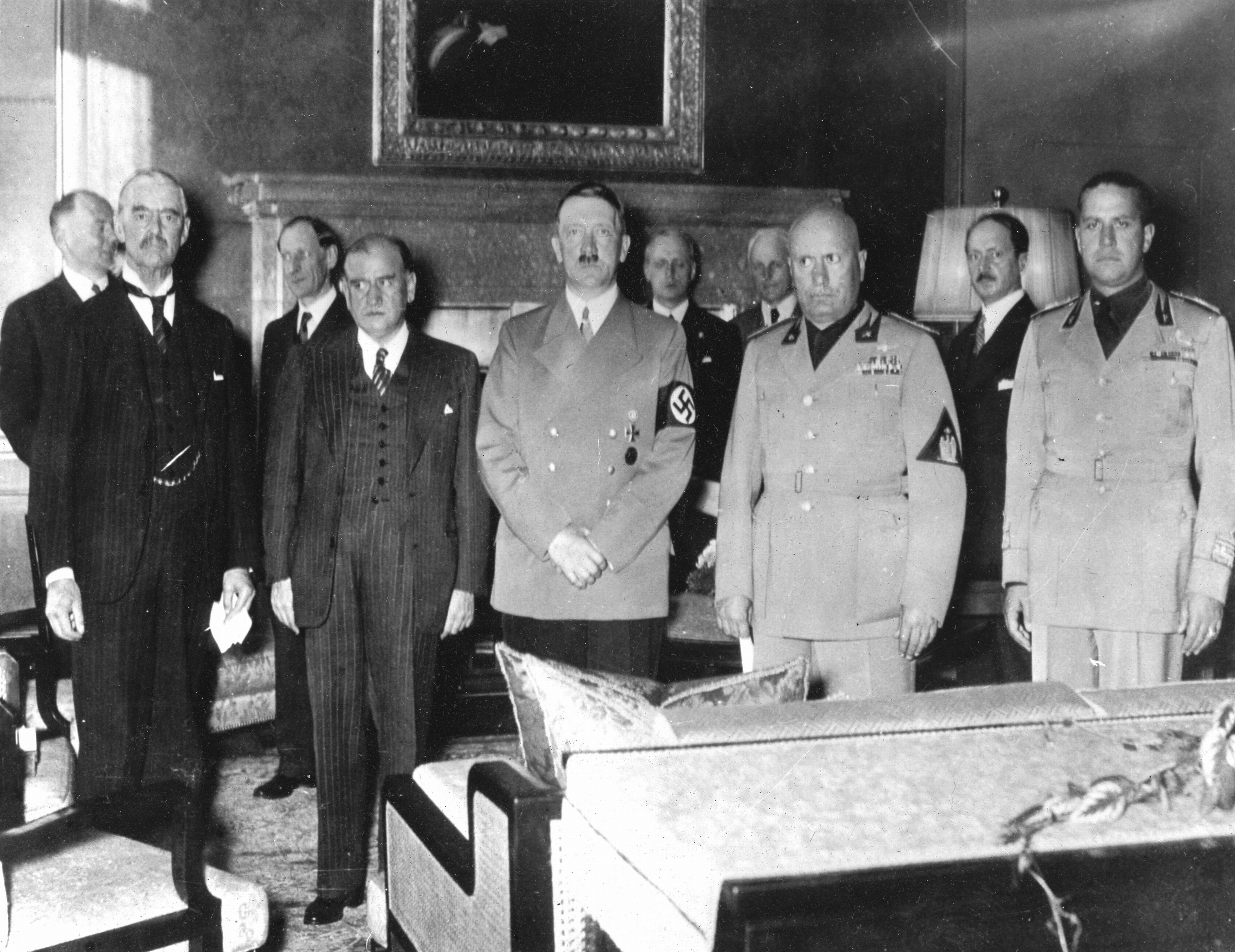 A group of men in formal and military attire, including Adolf Hitler with a Nazi arm cuff, stand in a room with a fireplace and large painting