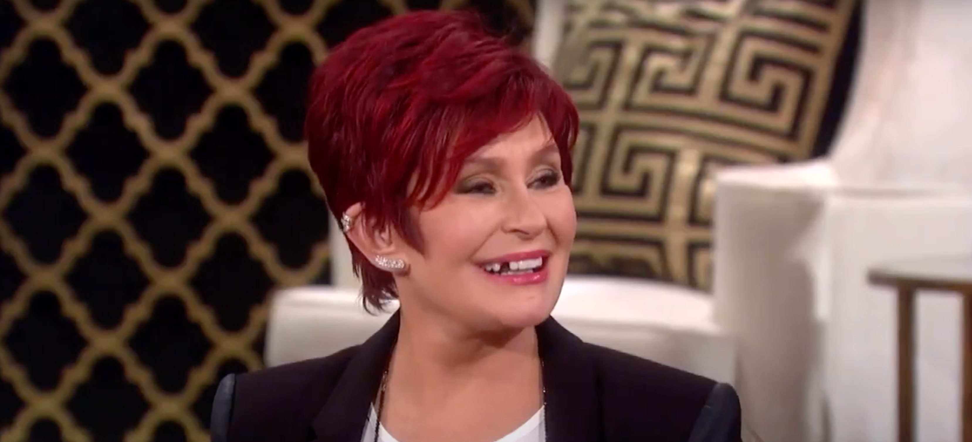 Person with short hair smiling, seated in a room with patterned cushions in the background. They're wearing a stylish black blazer over a white shirt