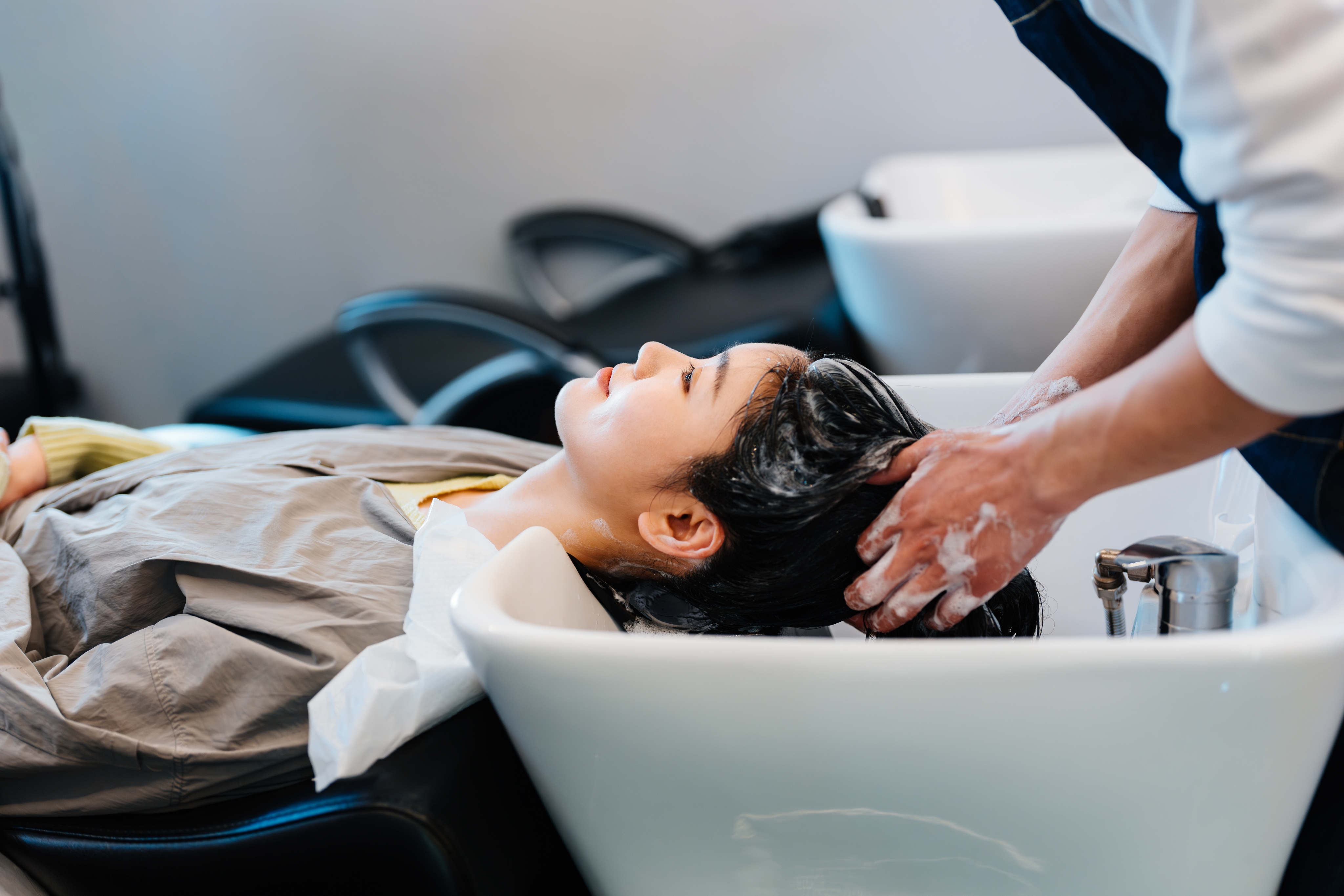 Person getting a hair wash at a salon, reclining with closed eyes, as a stylist massages shampoo into their hair at a washing station