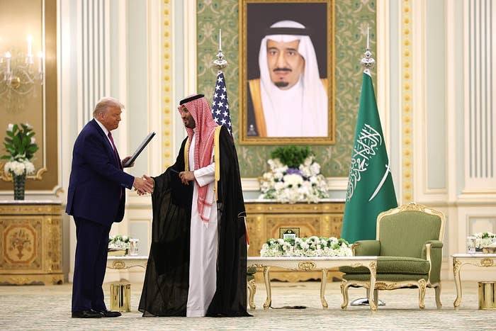 Two leaders shake hands in a formal setting with flags behind them. A portrait of a leader hangs on the wall