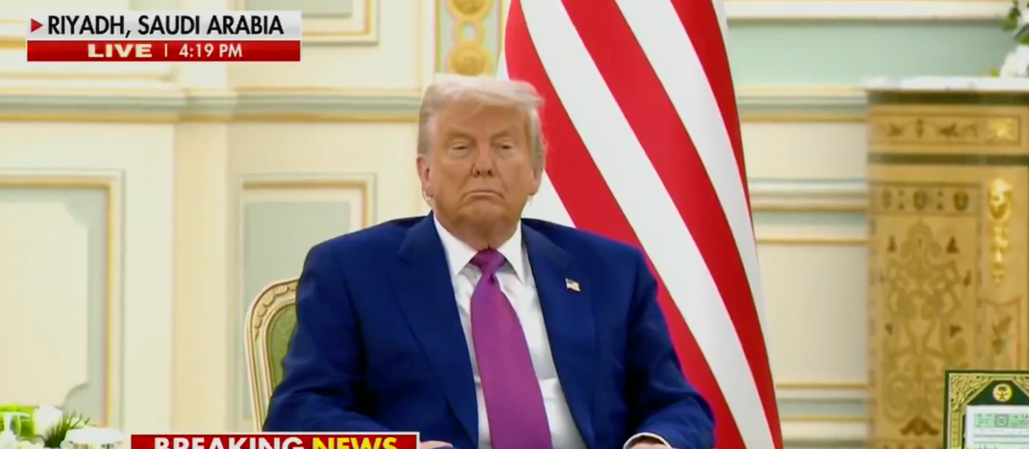 A person in a suit and tie sits in a formal setting, with flags and news ticker showing "Riyadh, Saudi Arabia" and "Breaking News."