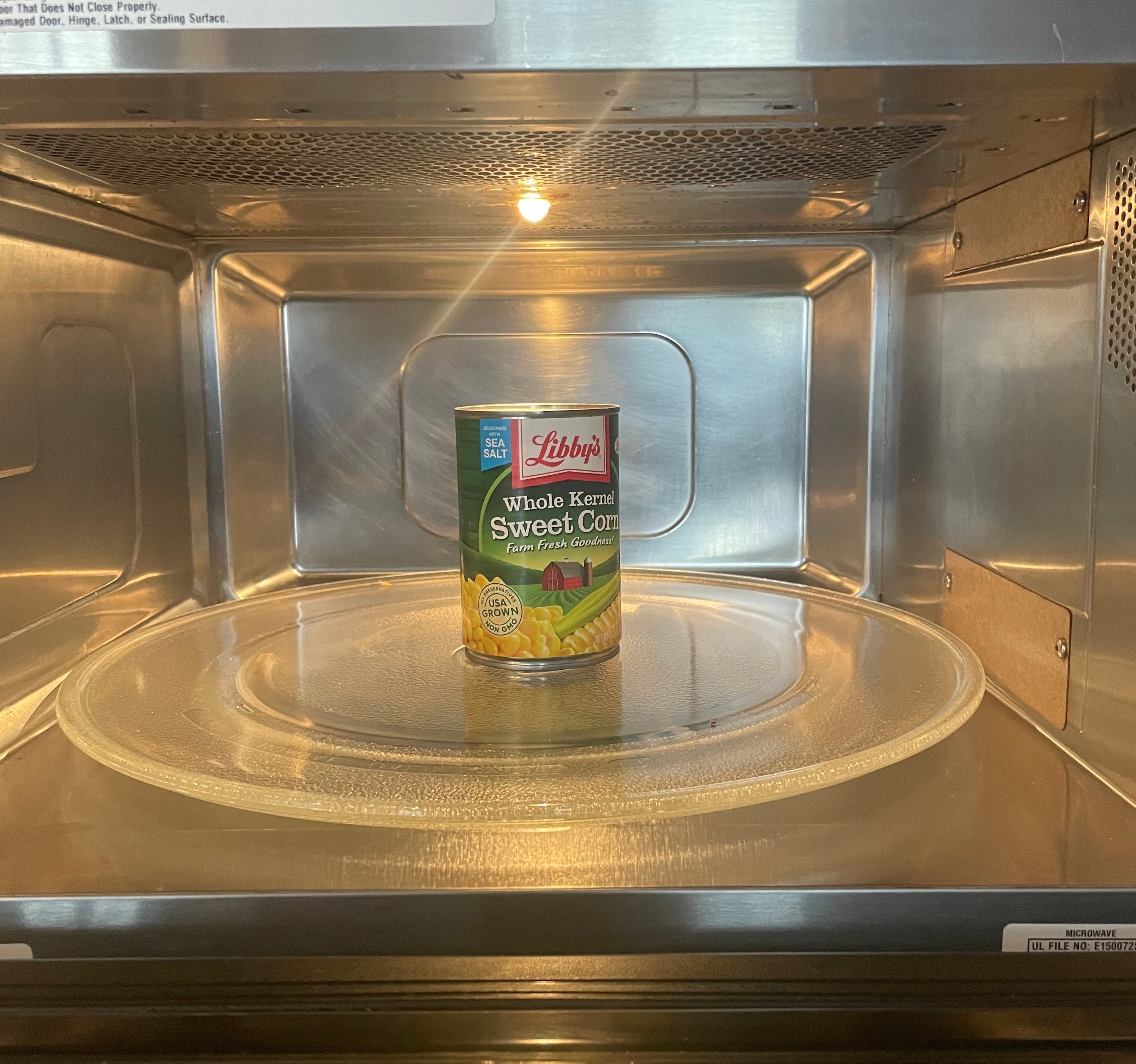 A can of sweet corn is sitting unopened inside a microwave, placed on the glass turntable