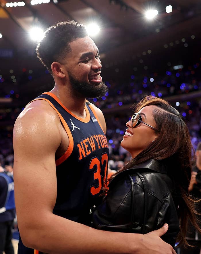 Basketball player in jersey embraces woman in sunglasses and leather jacket at an event. Both are smiling and appear happy