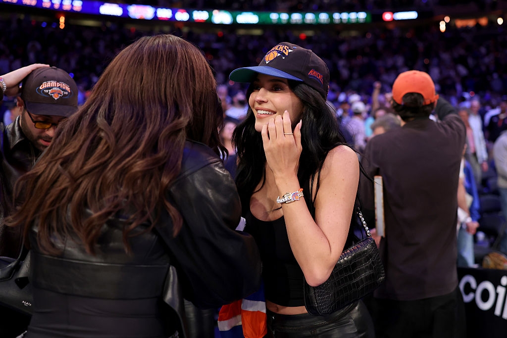Two women interact warmly at a basketball game, one wearing a casual cap and sleeveless top, suggesting a lively and friendly atmosphere