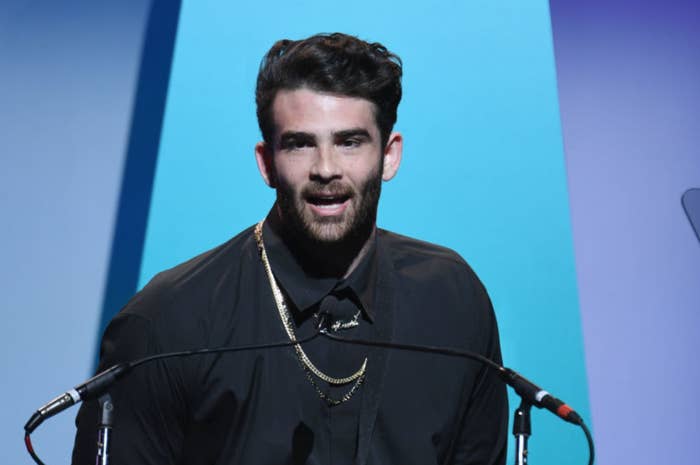 Man with dark hair and beard speaking at a podium, wearing a black shirt and gold chains