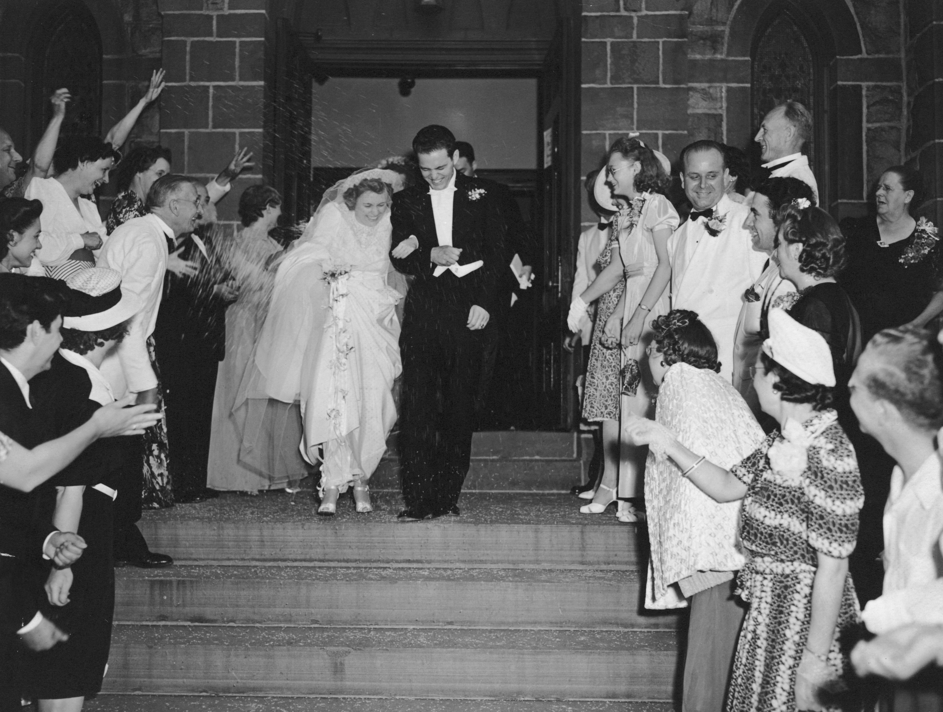 Bride and groom exit a church, dressed in traditional wedding attire, as guests cheer and throw rice in celebration