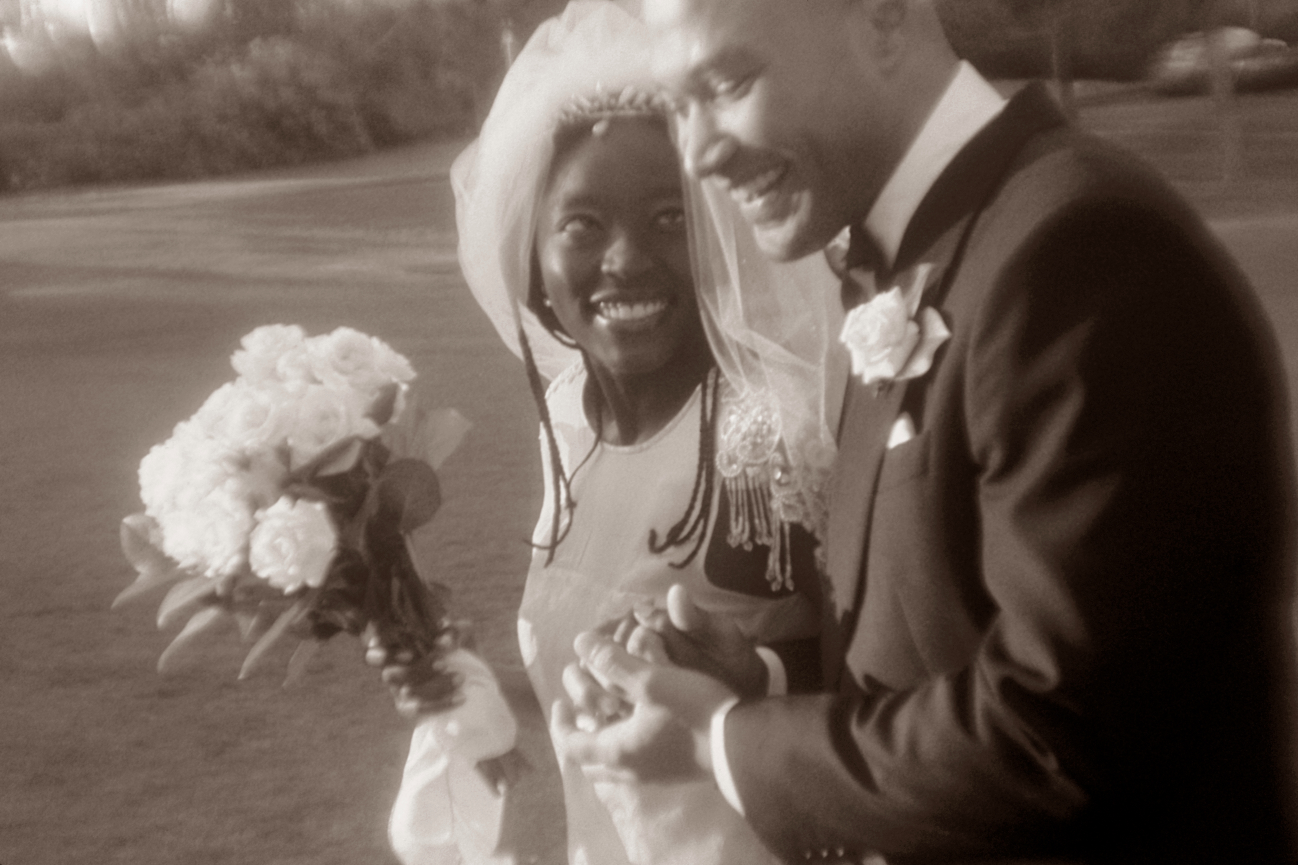 Bride in a flowing veil and elegant gown smiles at groom in a classic suit. Both hold hands, exuding joy as they celebrate their wedding outdoors