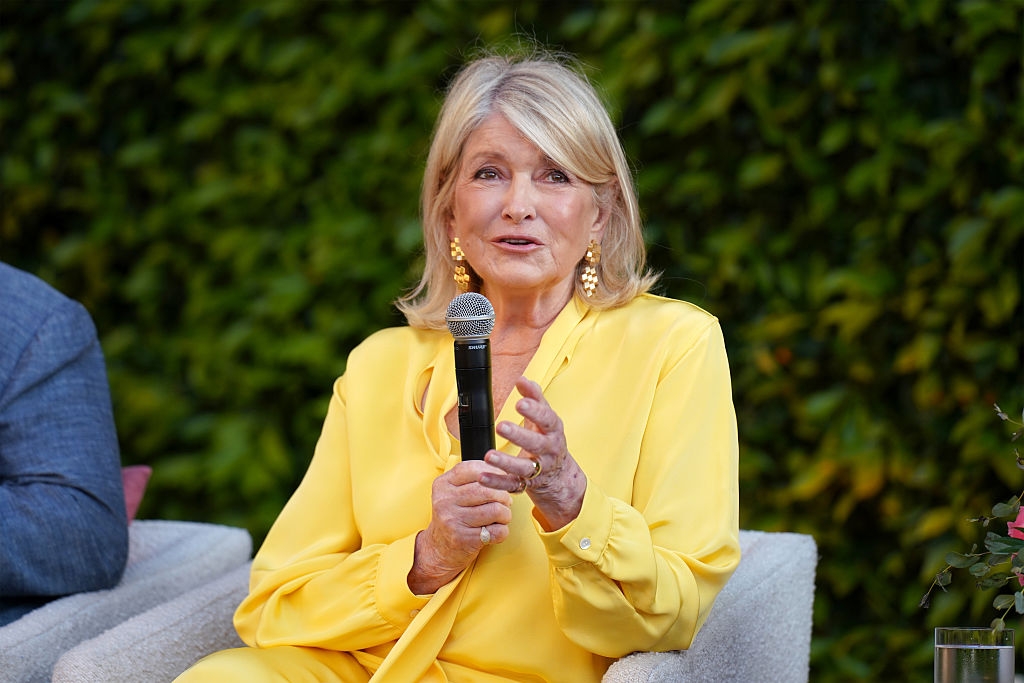 A woman in a stylish yellow outfit sits outdoors, holding a microphone, possibly speaking at an event