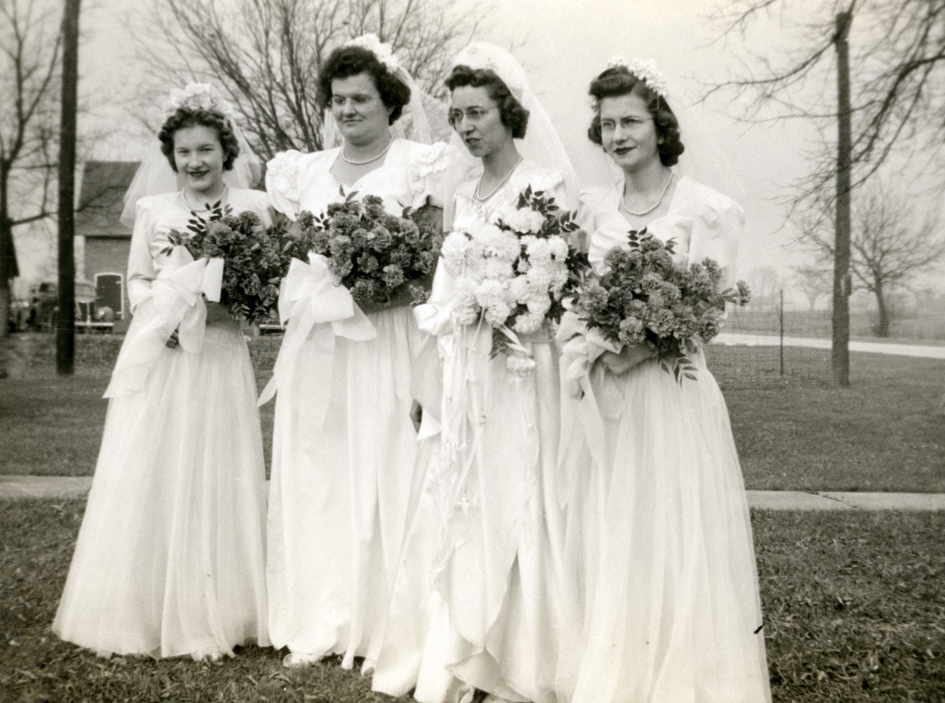 Four women in vintage bridal gowns hold bouquets outdoors. They are posing together, likely for a wedding photo, in a rural setting