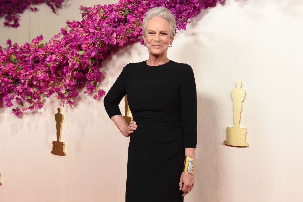 A person in a sleek black dress with an elegant updo poses confidently on a red carpet, surrounded by floral decorations and Oscar statuette images