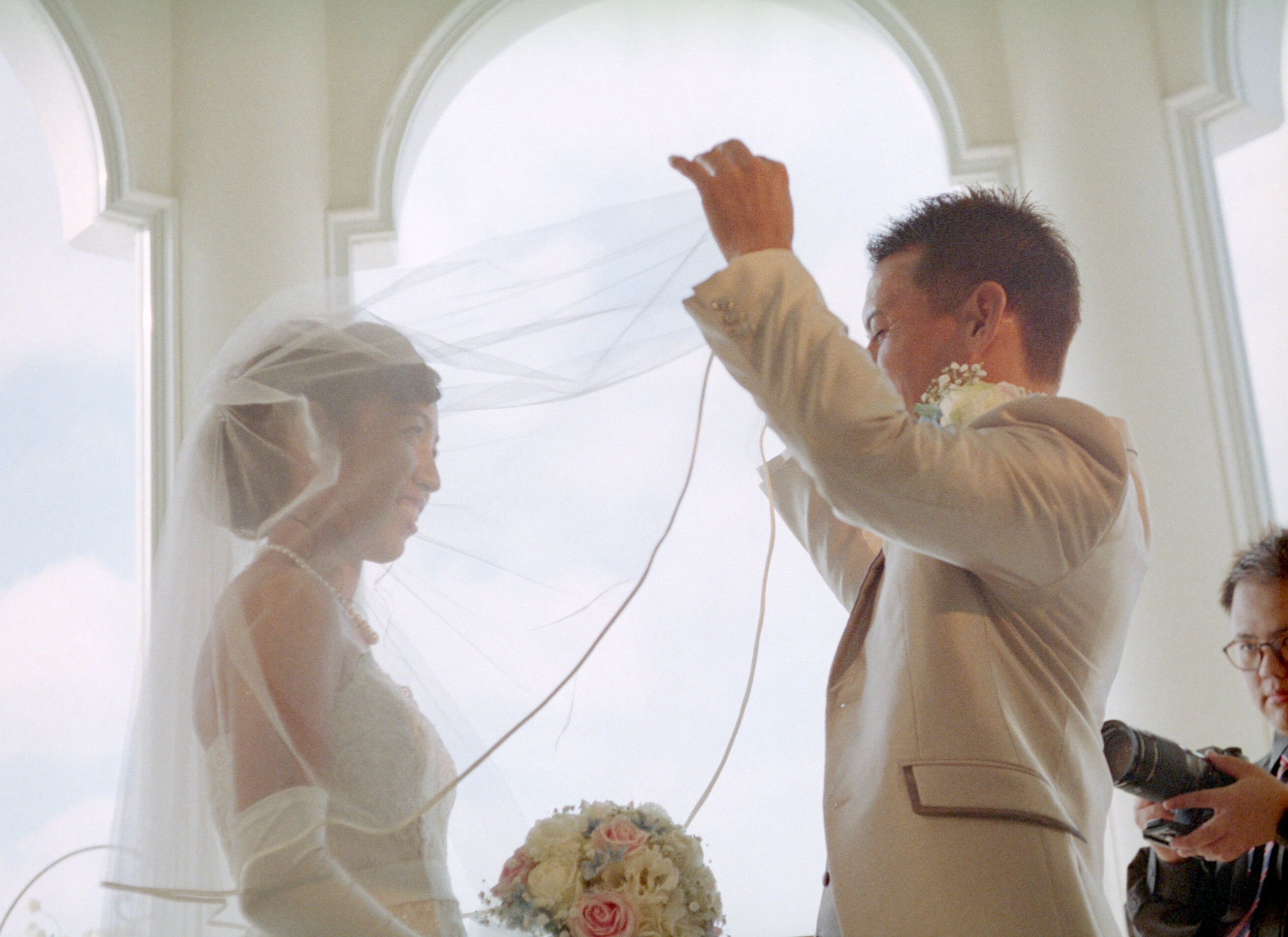 Couple at wedding ceremony; groom lifts bride's veil. Bride in elegant gown, groom in light suit. Romantic setting with arched windows in background