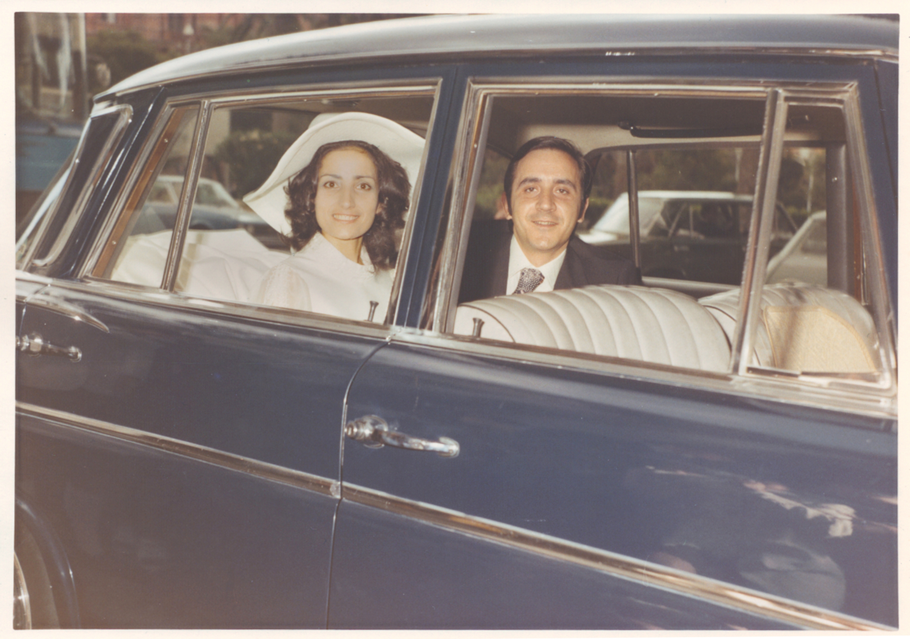 Bride and groom smile from a classic car's backseat, with the bride in a wide-brimmed hat and elegant attire, celebrating their wedding day