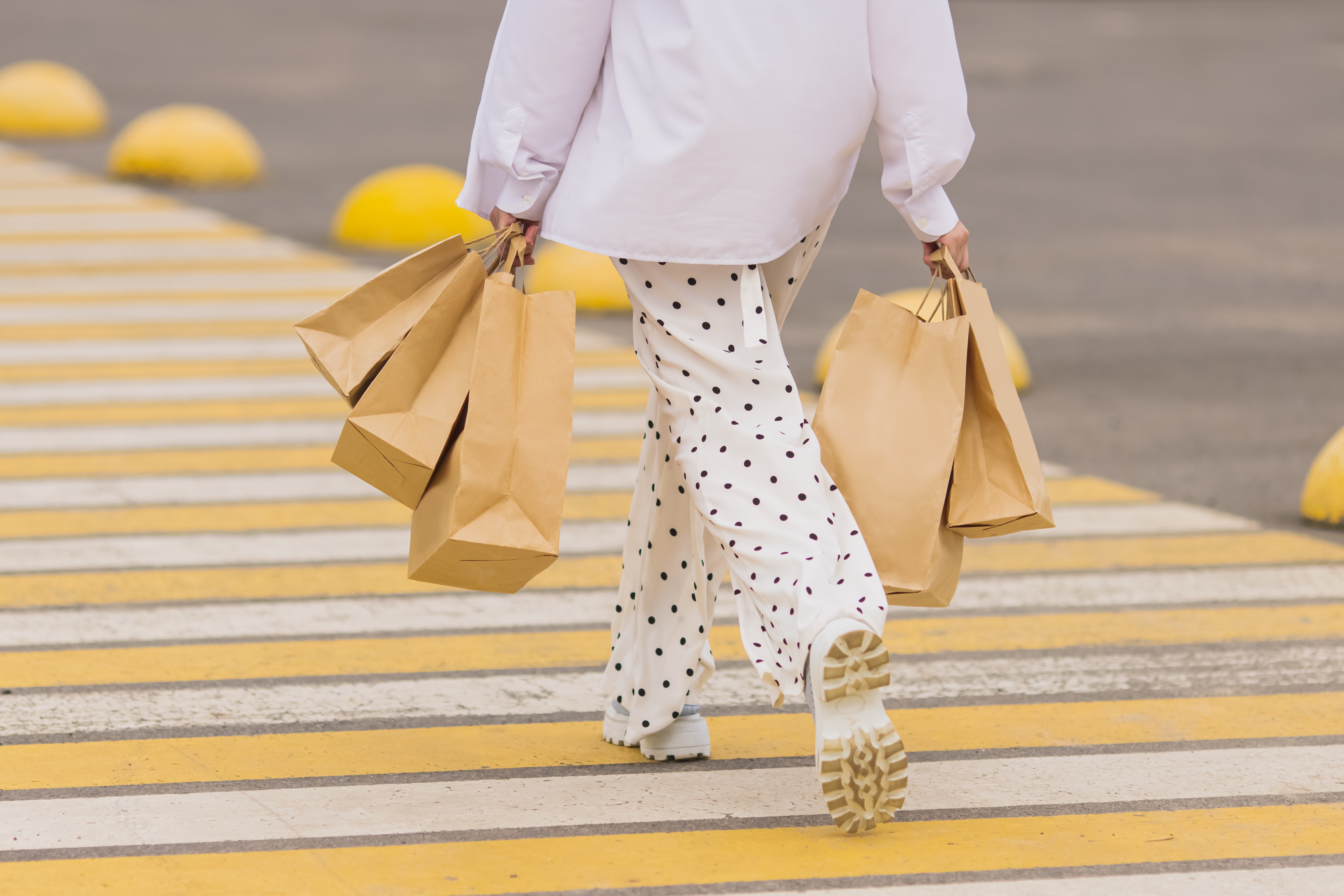 Person in polka dot pants carrying shopping bags crosses a street with yellow-striped crosswalk