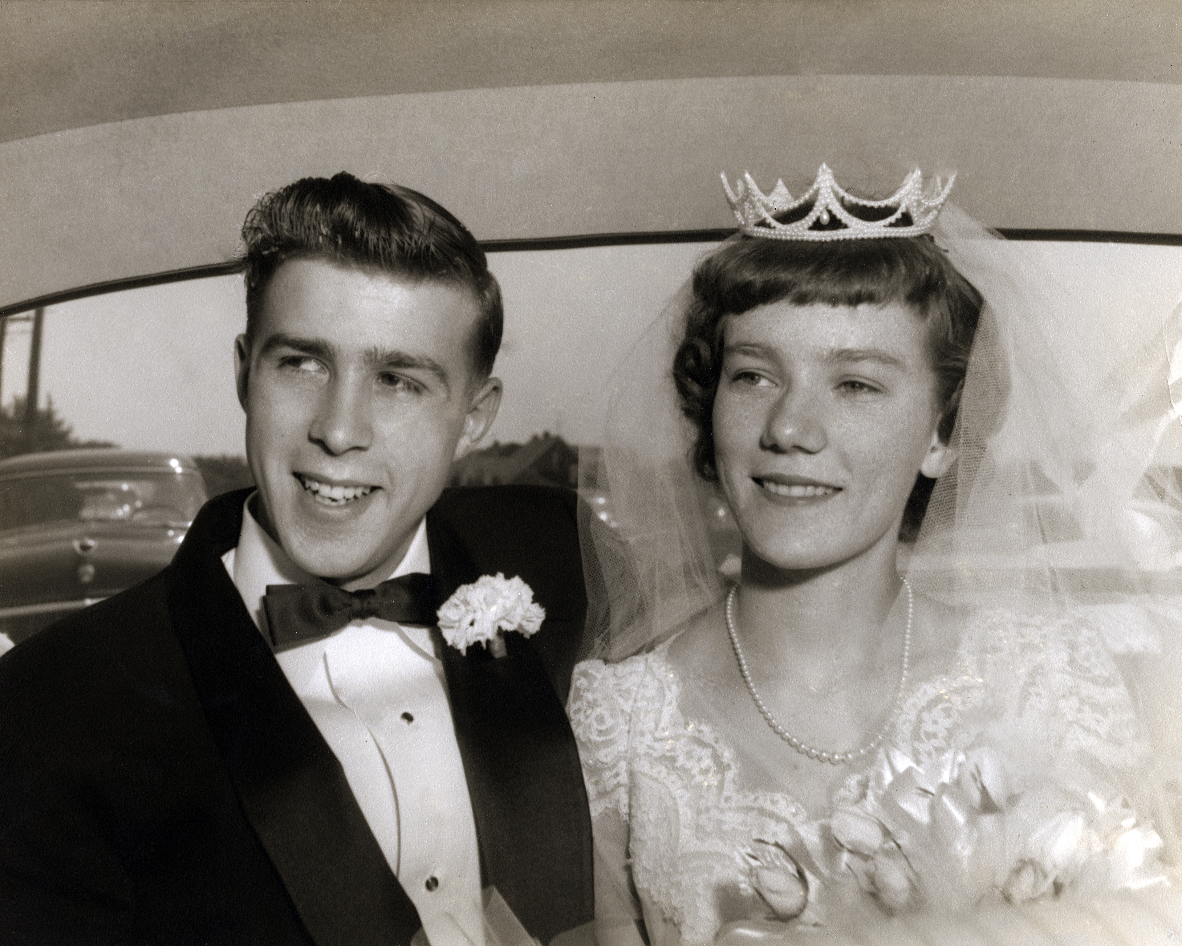 A couple in a vintage wedding photo; the groom wears a tuxedo with a bow tie, and the bride wears a lace dress with a tiara and veil