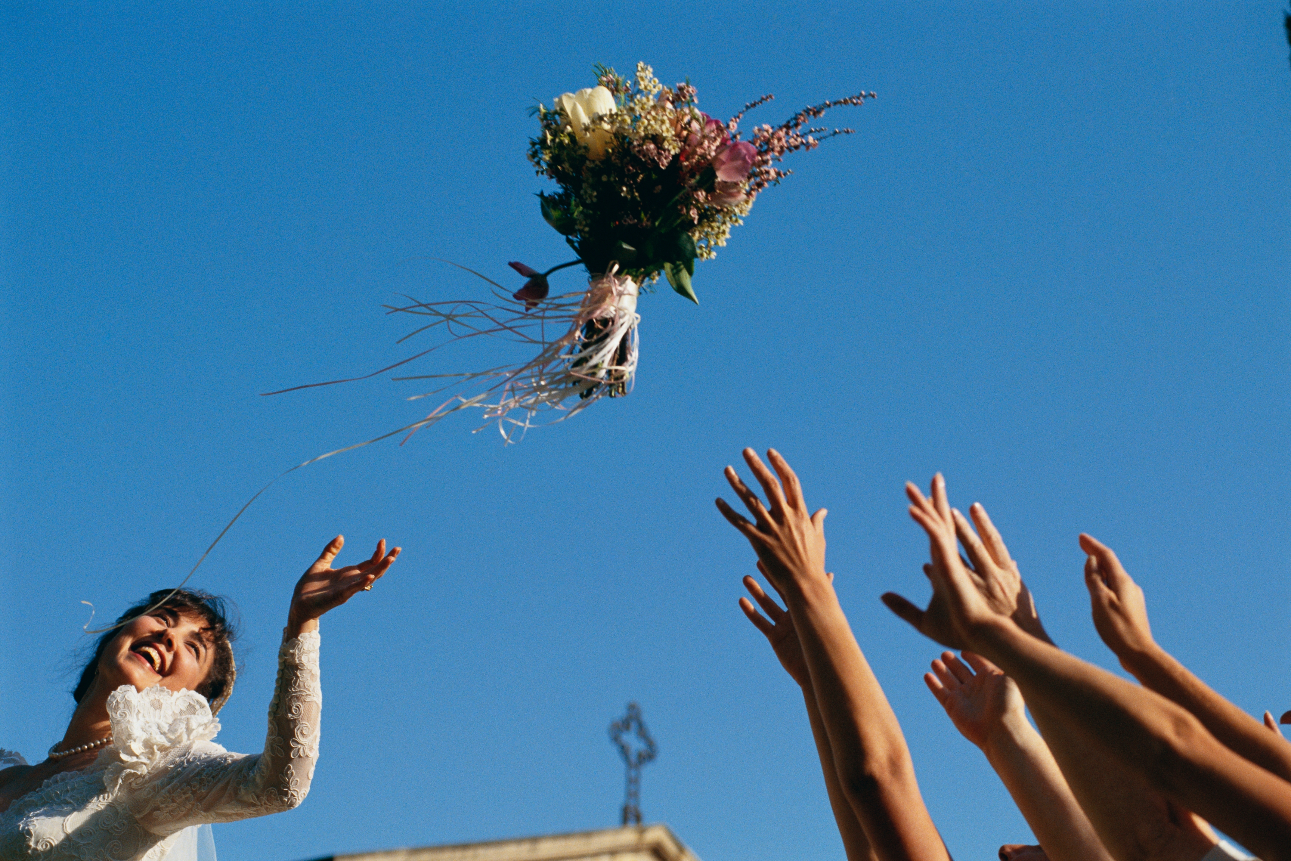 Bride joyfully tosses bouquet into air, with guests reaching out to catch it, set against a clear sky during a wedding celebration
