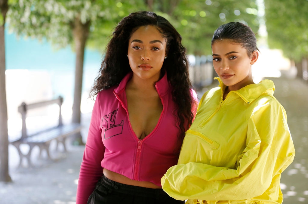 Two individuals stand outdoors, one in a bright pink zip-up with long curls, and the other in a yellow jacket with hair pulled back