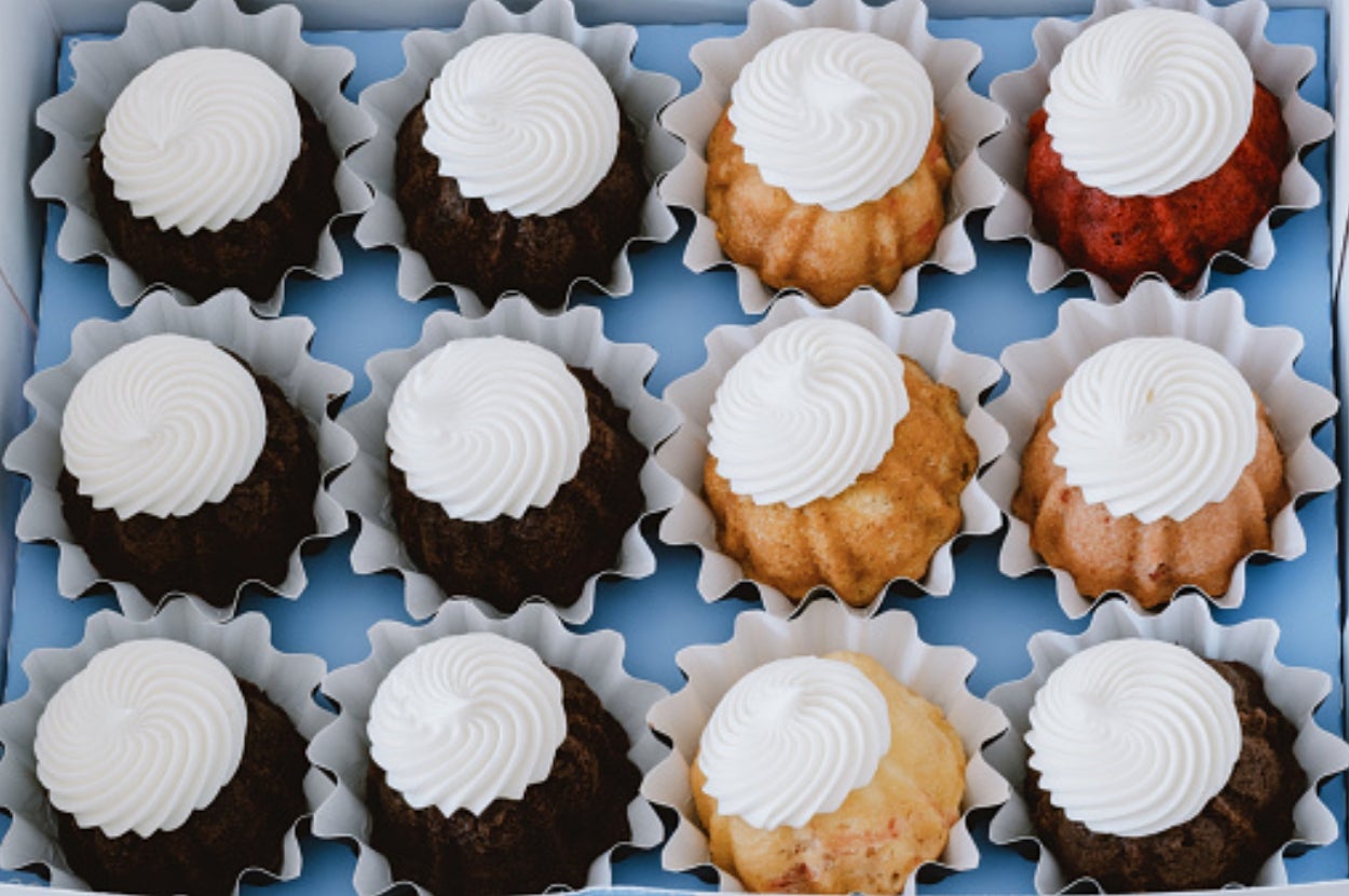 A box of twelve small bundt cakes with swirl frosting, each in a paper cup, displayed in a grid pattern