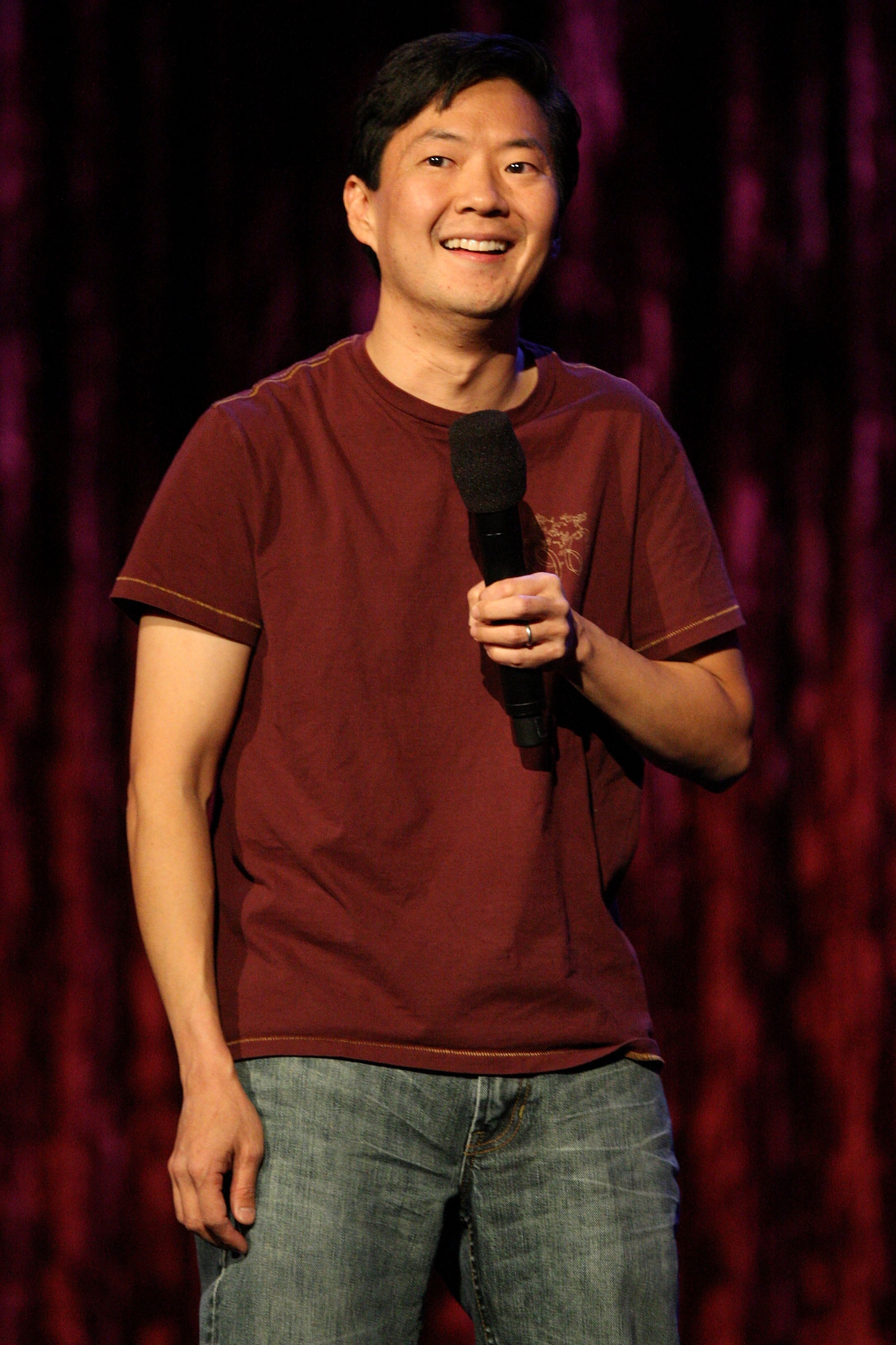 Ken Jeong holding a microphone, wearing a casual t-shirt and jeans, smiling while standing in front of a curtain backdrop