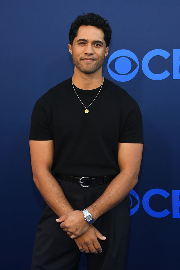 Man on blue background wears a black t-shirt, dark pants, belt, and a necklace, posing calmly