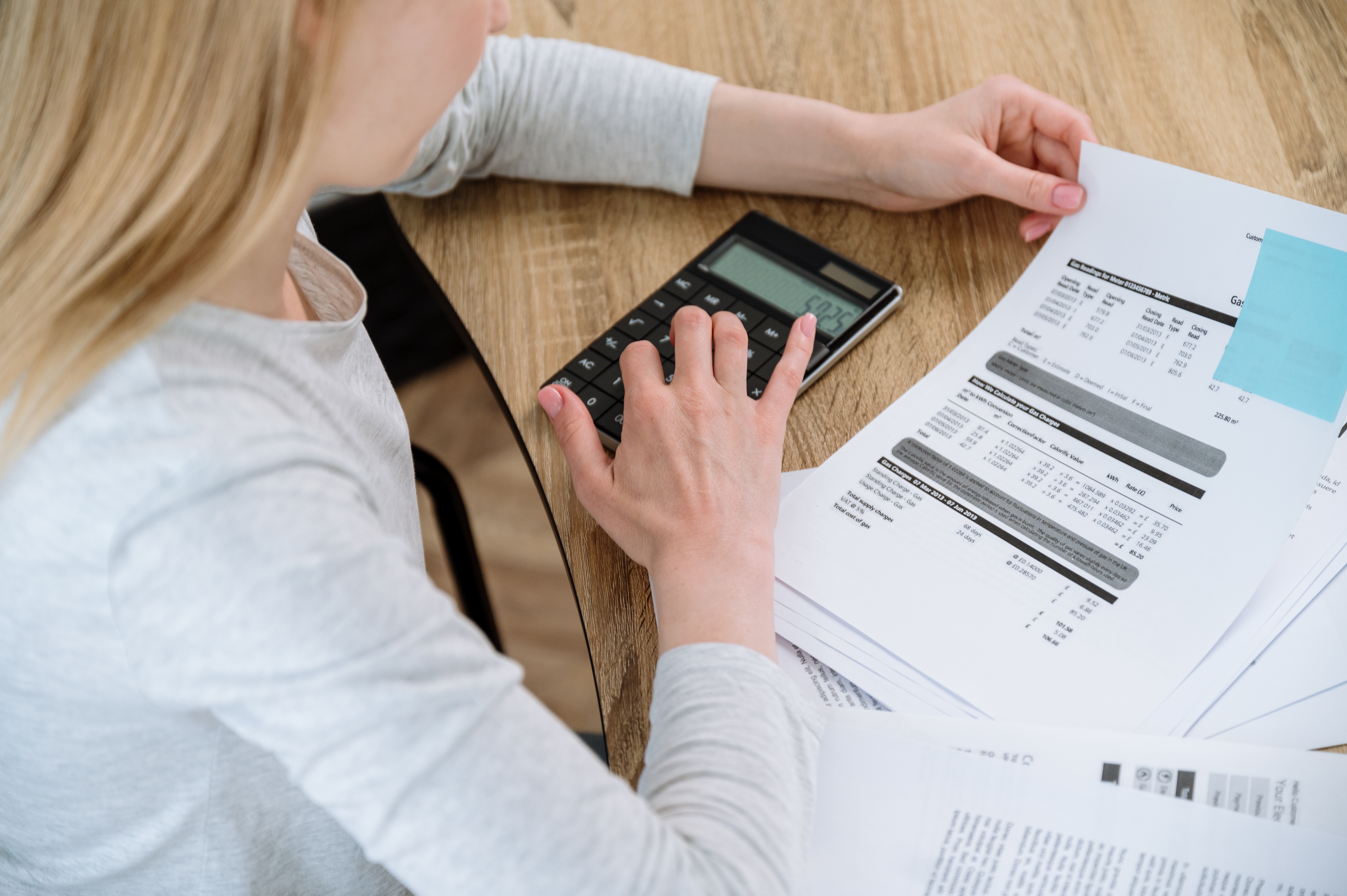 Person using a calculator at a wooden table, reviewing financial documents and printed pages
