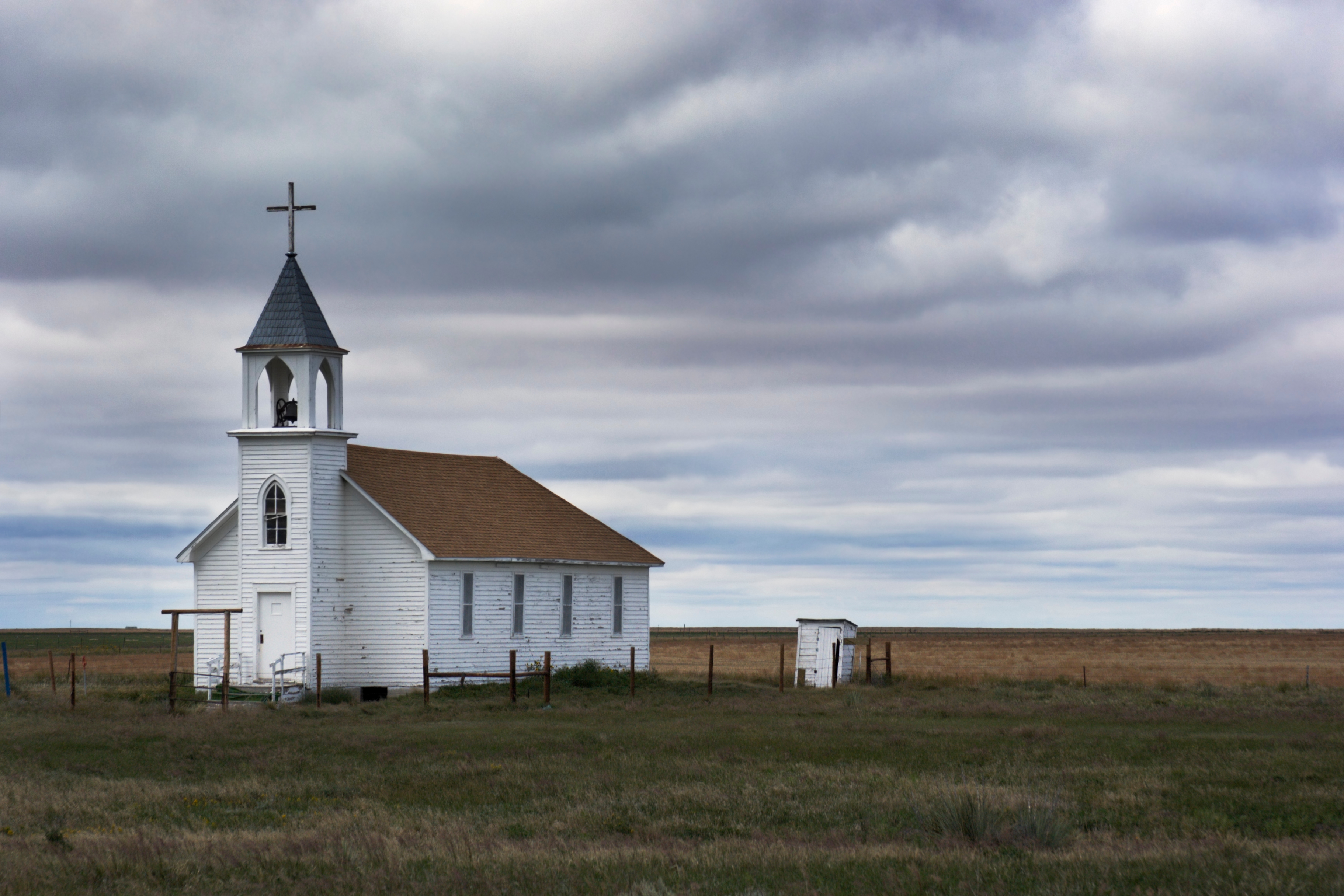 A small white church with a steeple stands alone in a vast open field under a cloudy sky