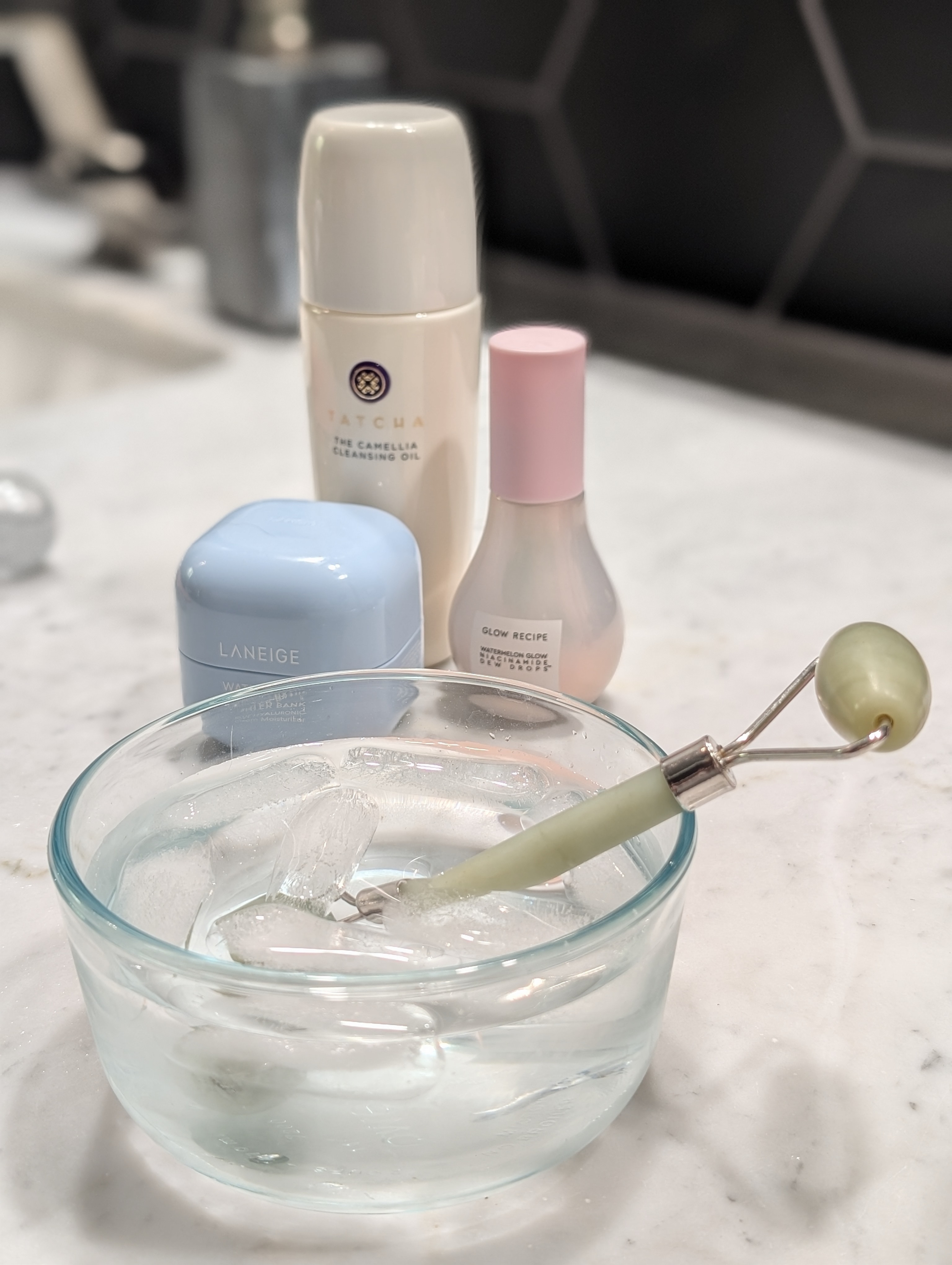 Various skincare products on a marble countertop with a jade roller in a bowl of ice water, suggesting a skincare routine
