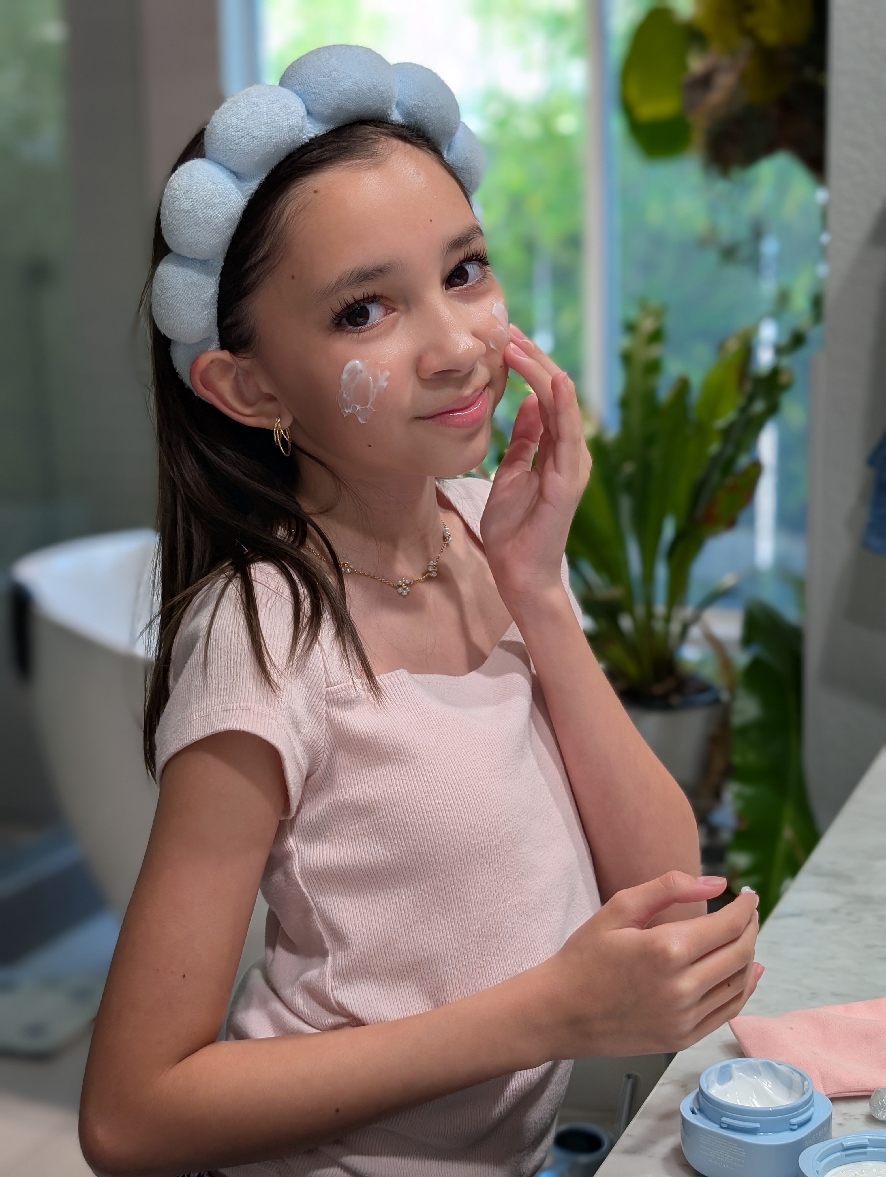 Young girl applying face cream, wearing headband and gold earrings, in a bathroom setting