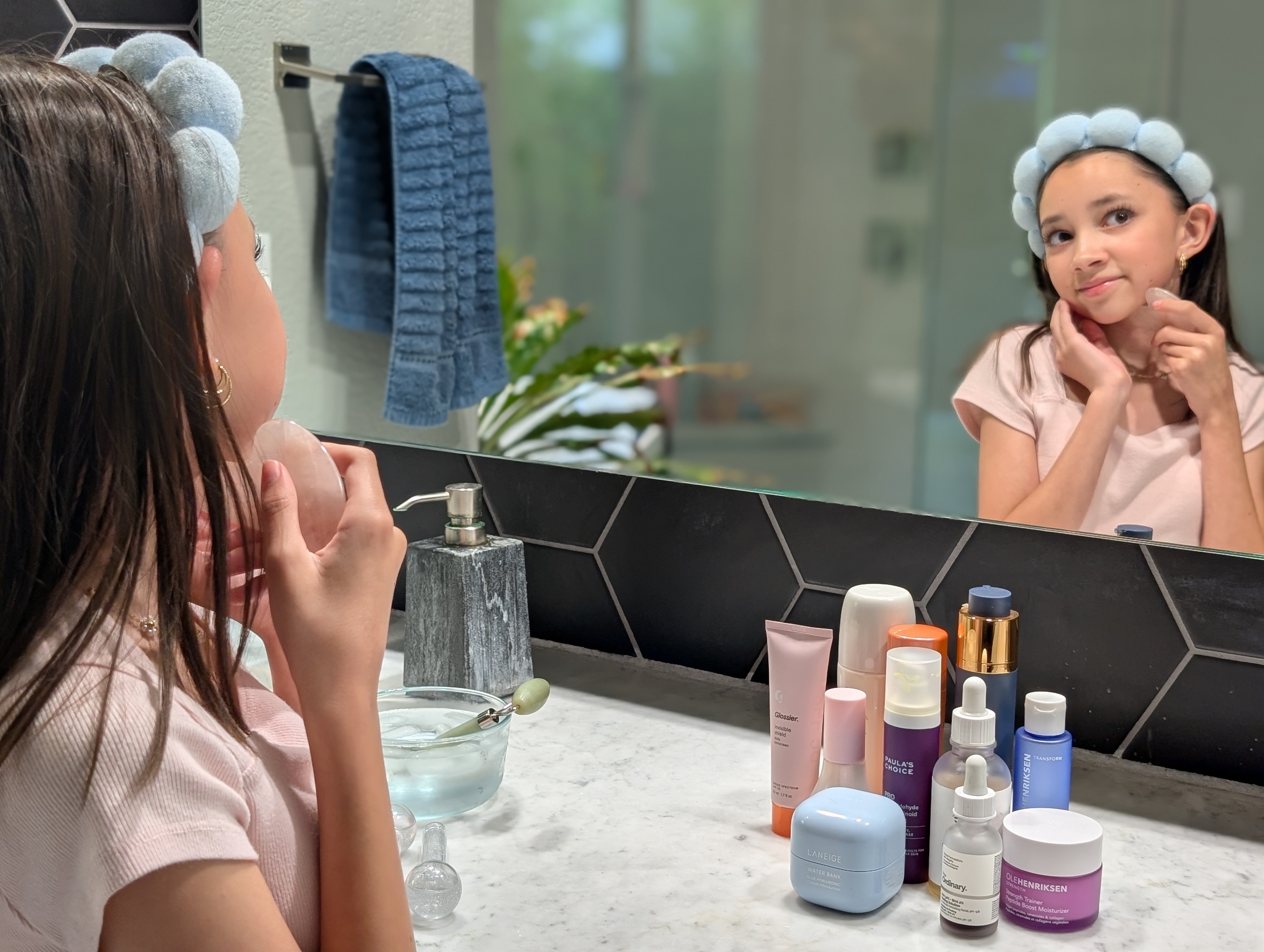 Child with headband applies skincare in bathroom mirror, surrounded by various products
