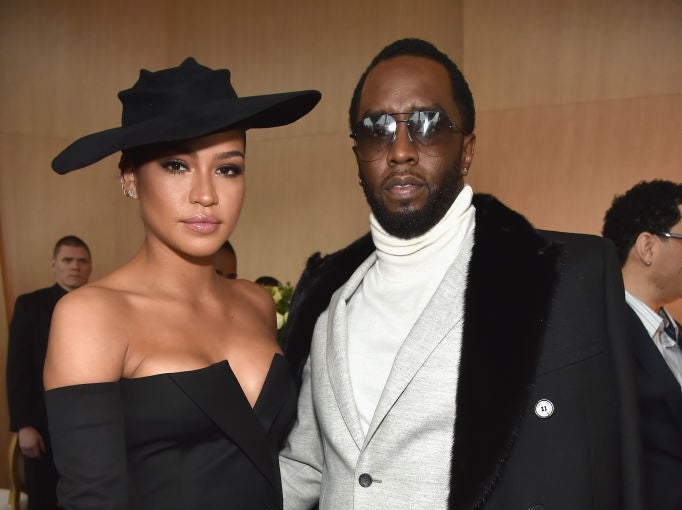 Two individuals pose together; one is wearing a stylish off-shoulder dress and hat, the other in a sleek suit with a fur-collared coat and turtleneck