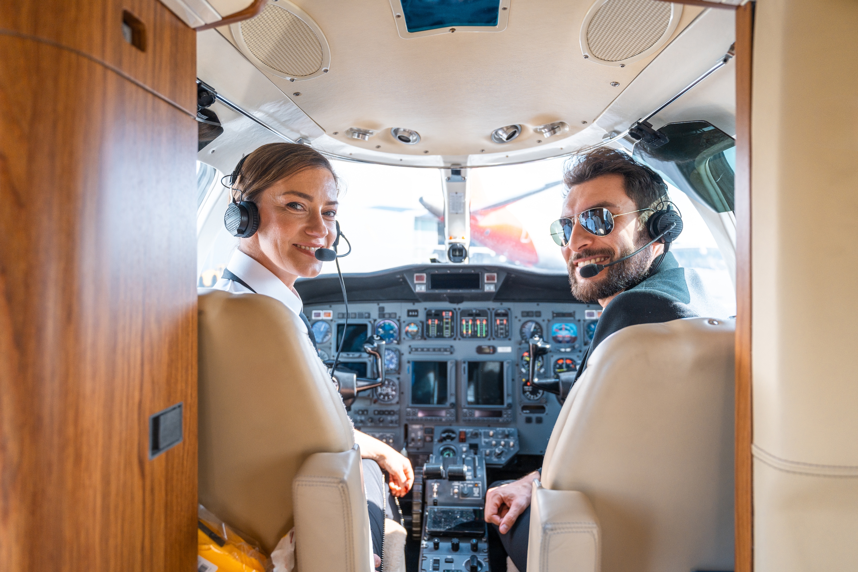 Two pilots wearing headsets smile from an airplane cockpit, looking towards the camera. The cockpit displays various flight controls