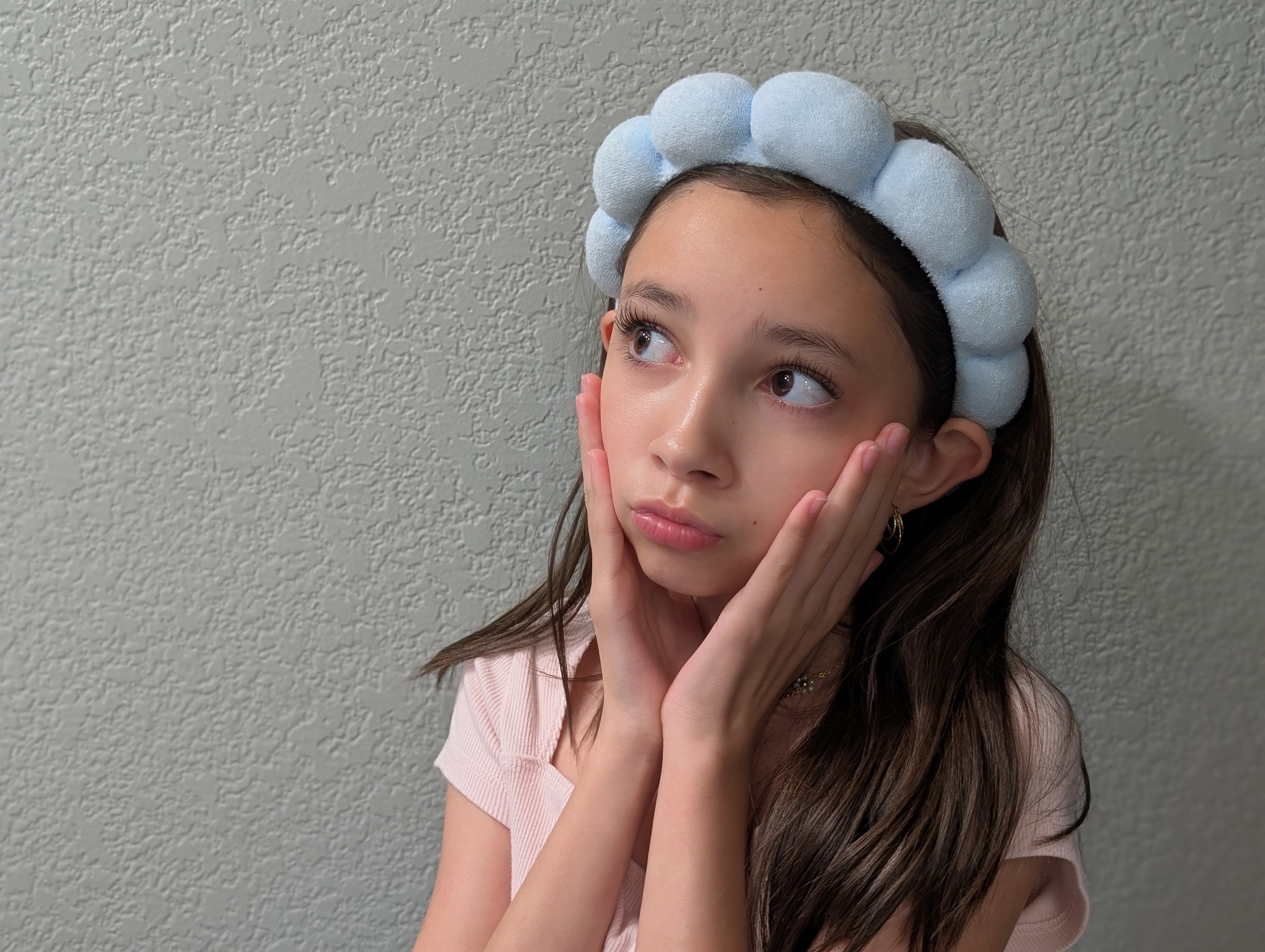 Child with a playful expression wearing a round bubble-like headband, hands on cheeks, looking thoughtfully upward against a plain background